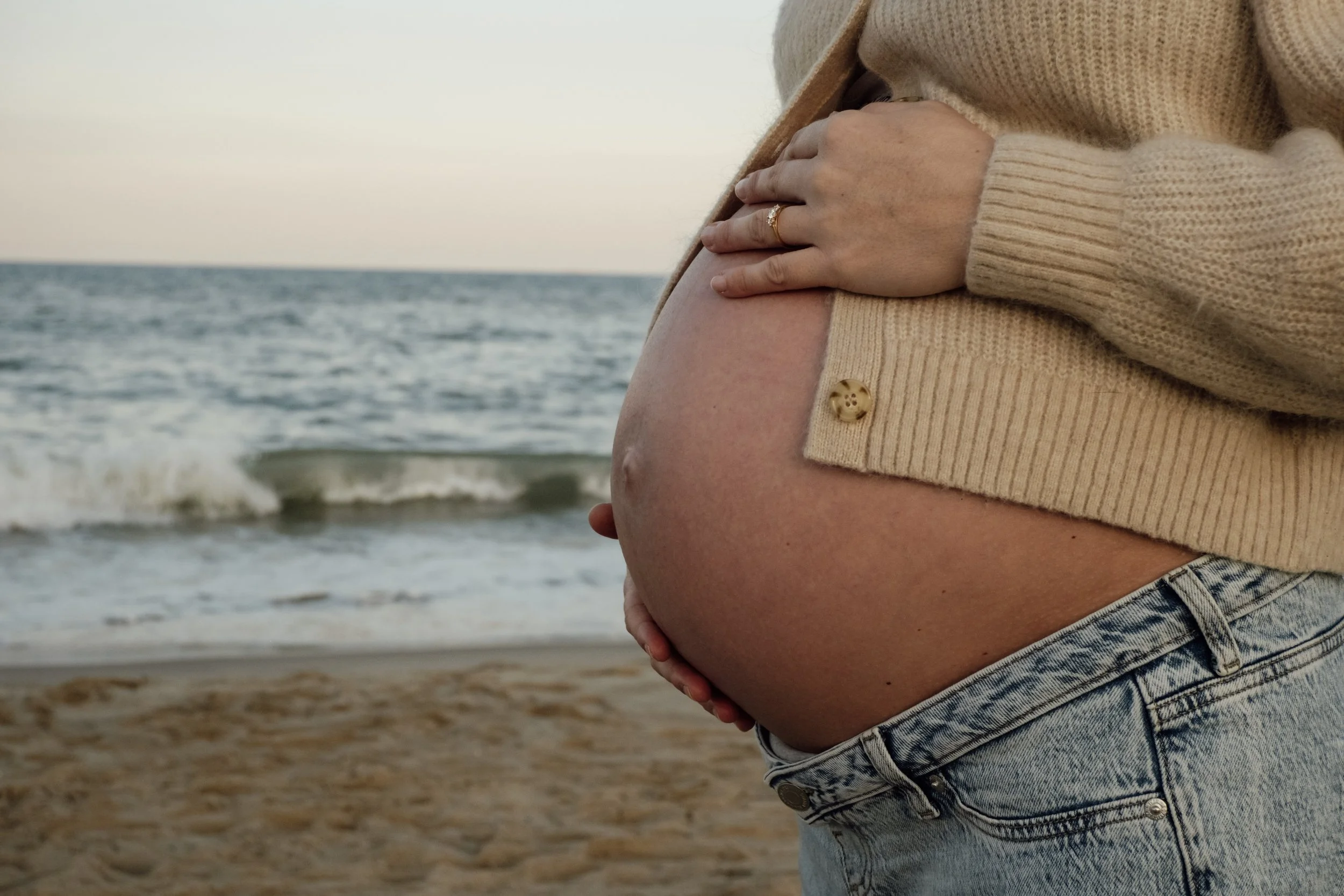 Pregnant woman on the beach holding her belly with both hands, wearing a beige sweater and denim shorts.