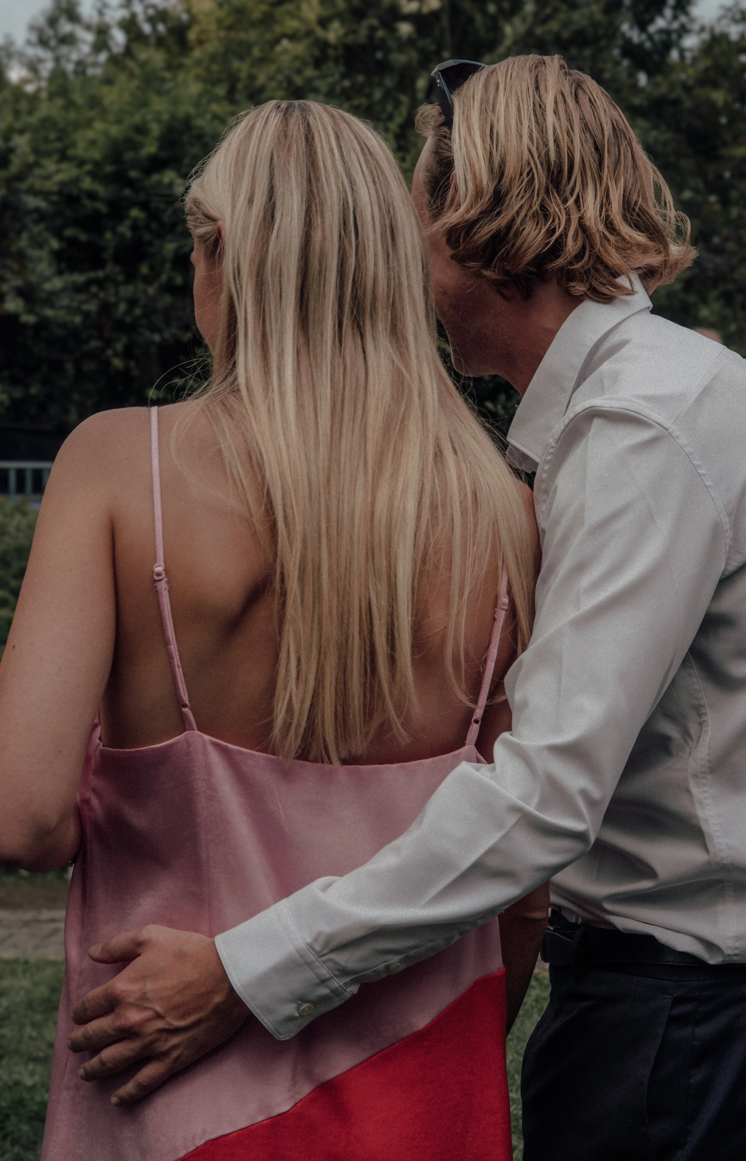 A man and a woman, both with blonde hair, are close together outdoors. The man is wearing a white shirt and glasses on his head. The woman is wearing a pink strap dress. They are sharing a kiss or whispering intimately.