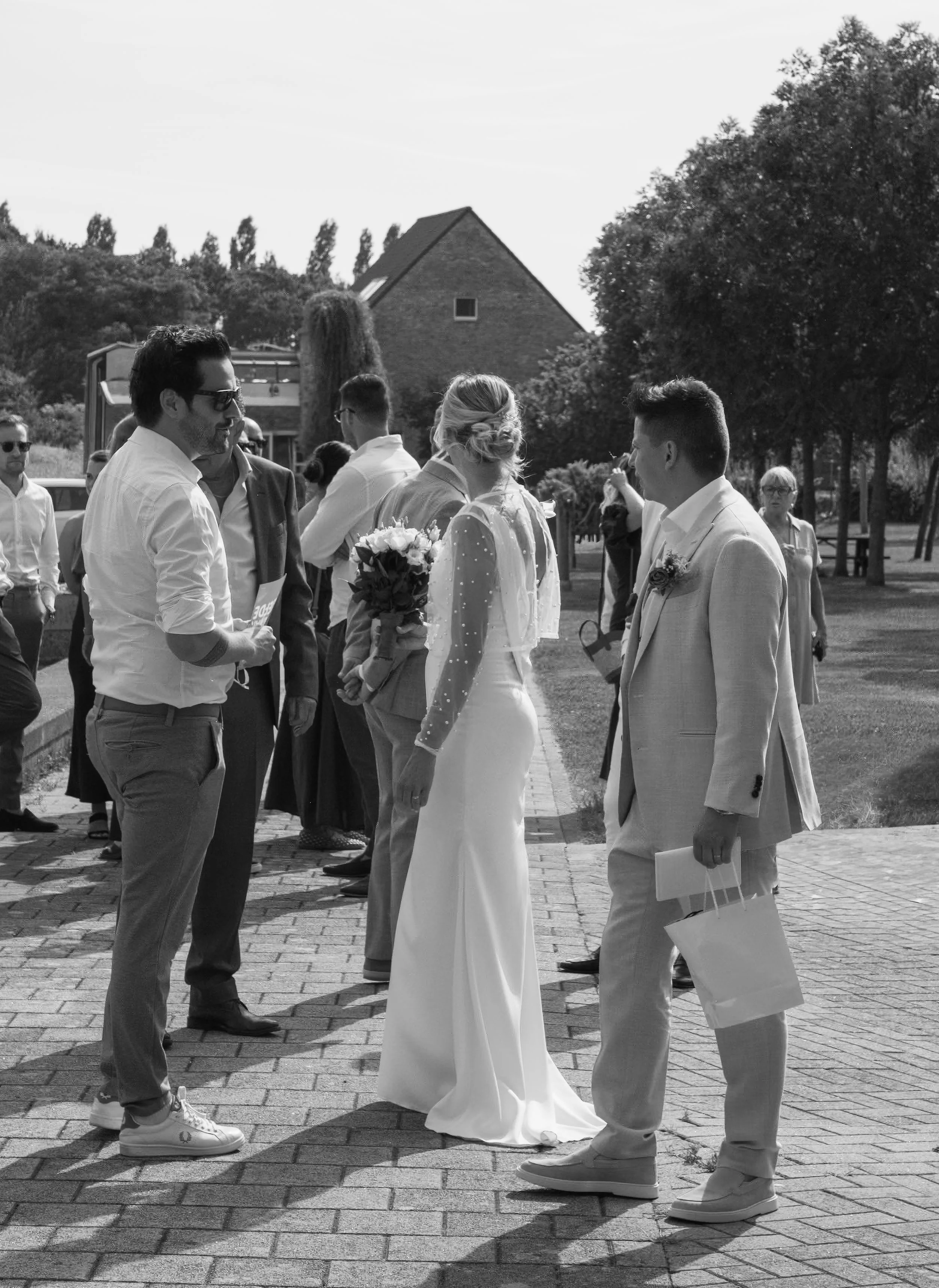 A wedding ceremony outdoors with a bride in a long gown holding a bouquet and three men in suits, two wearing white and one in a light-colored suit, standing on a paved walkway surrounded by trees and other guests.