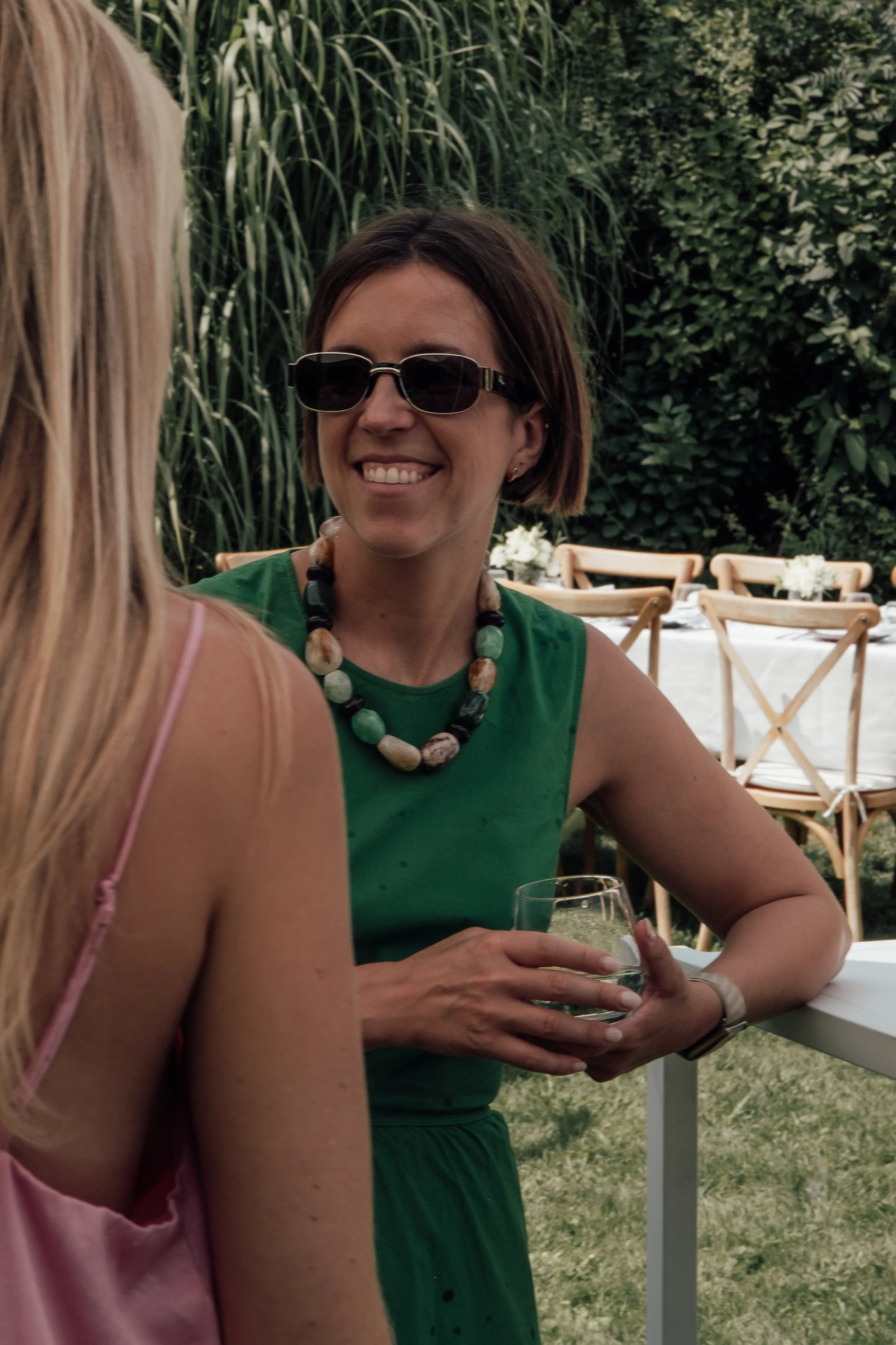 Two women having a conversation outdoors, one with blonde hair wearing a pink top and the other with dark hair wearing sunglasses and a green top, seated at a table with a glass of water, surrounded by greenery and outdoor furniture.