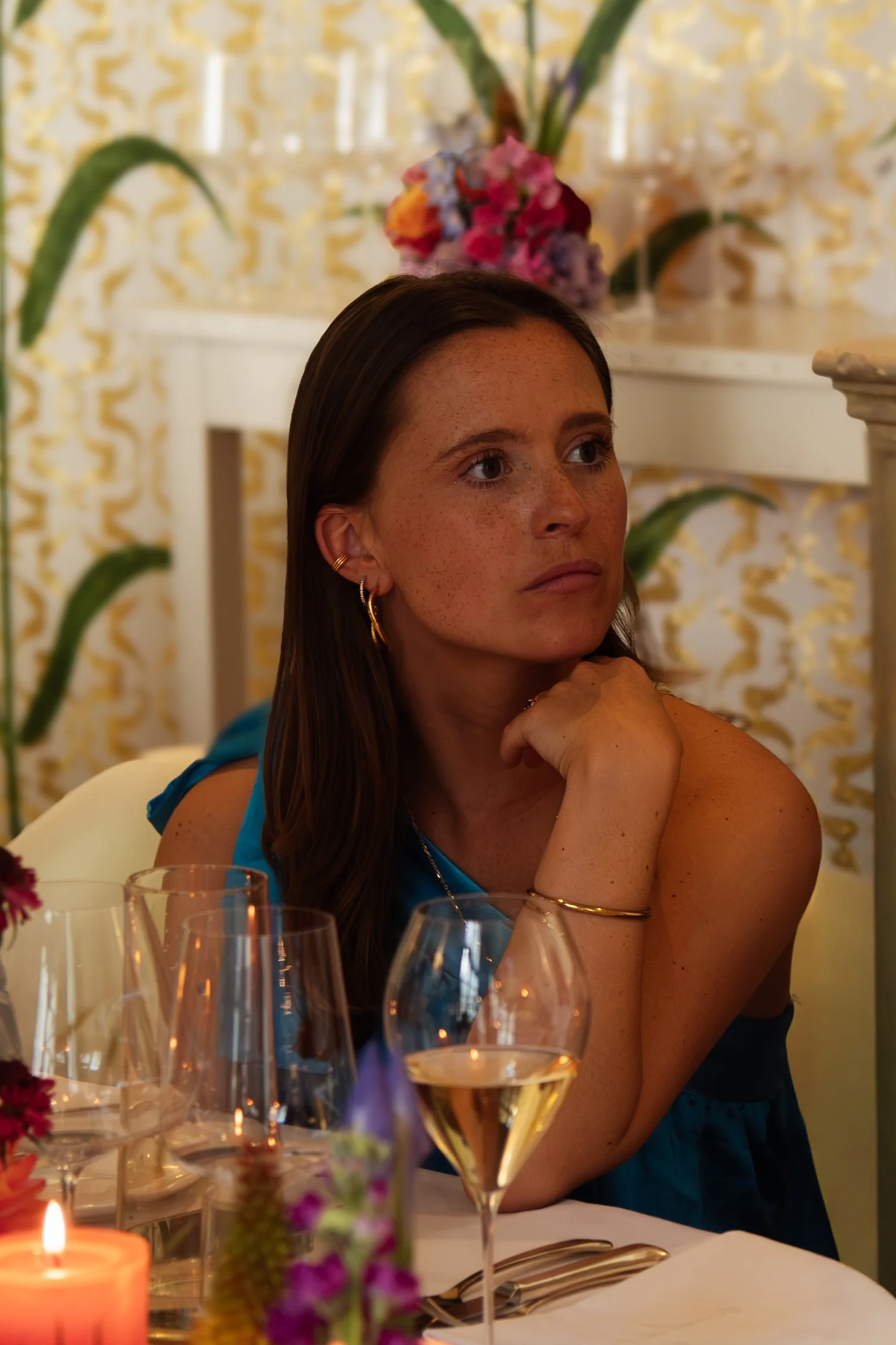 A woman with long brown hair, wearing earrings and a gold bracelet, is sitting at a decorated table with multiple glasses of wine, a candle, and floral decorations. She appears to be listening or contemplating, with her hand resting on her chin, in a