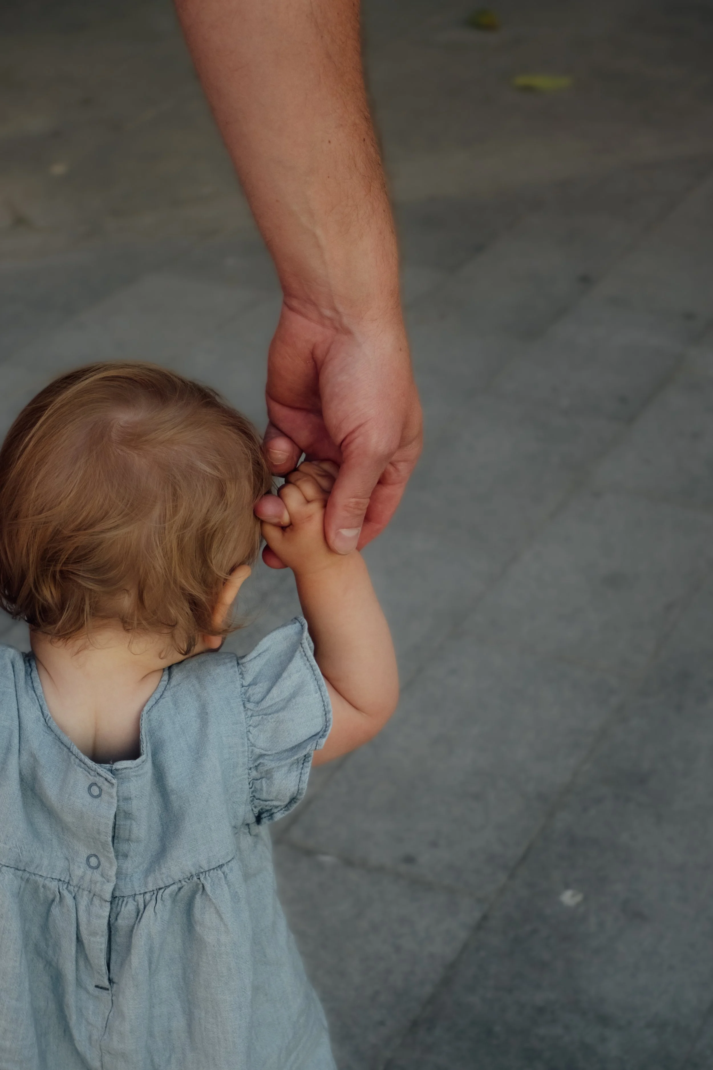 A young child with curly blond hair holding an adult's hand, walking on a paved outdoor surface.