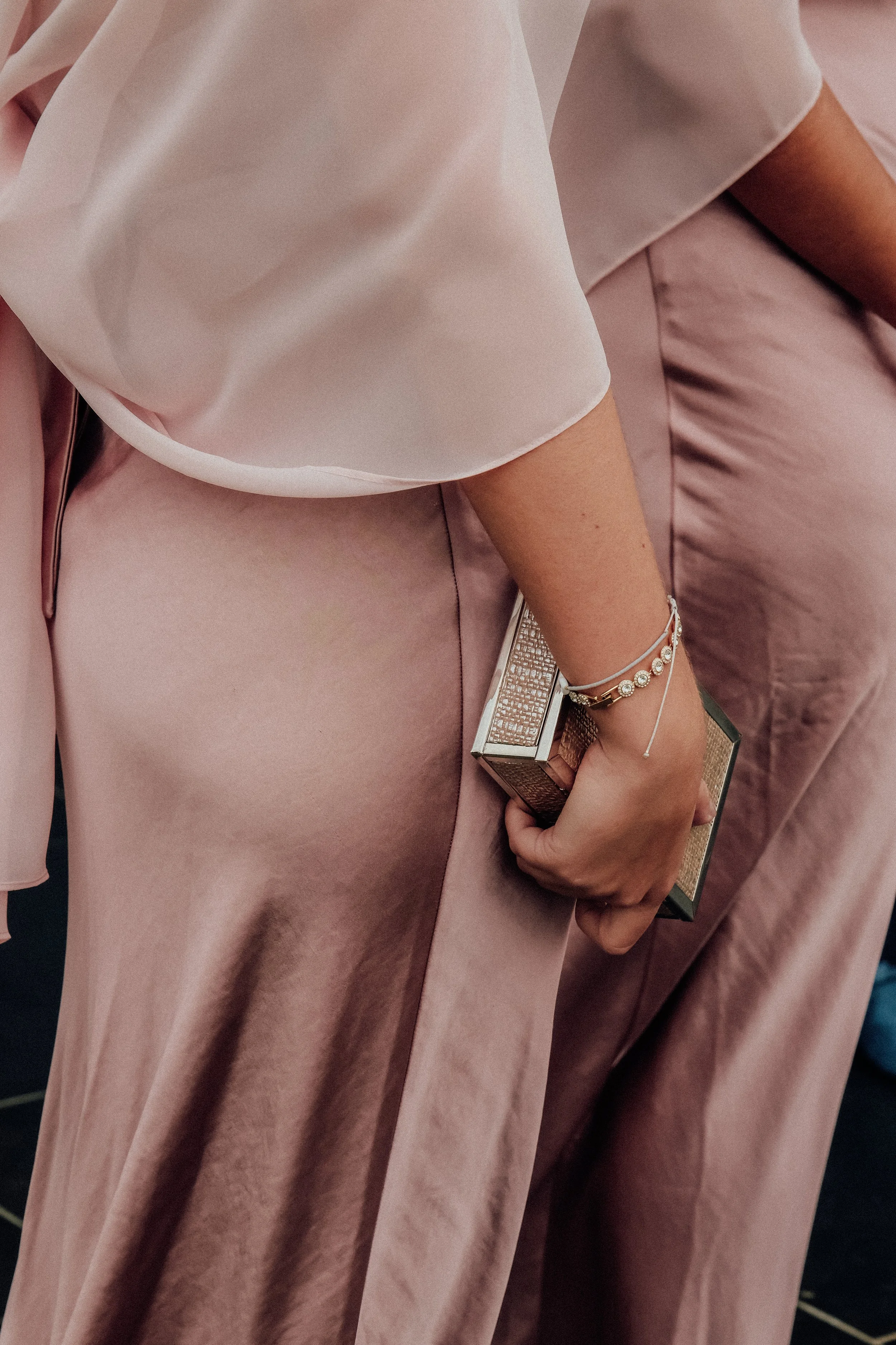 Close-up of a person wearing a pale pink satin dress and holding a glittering clutch purse, with jewelry on their wrist.