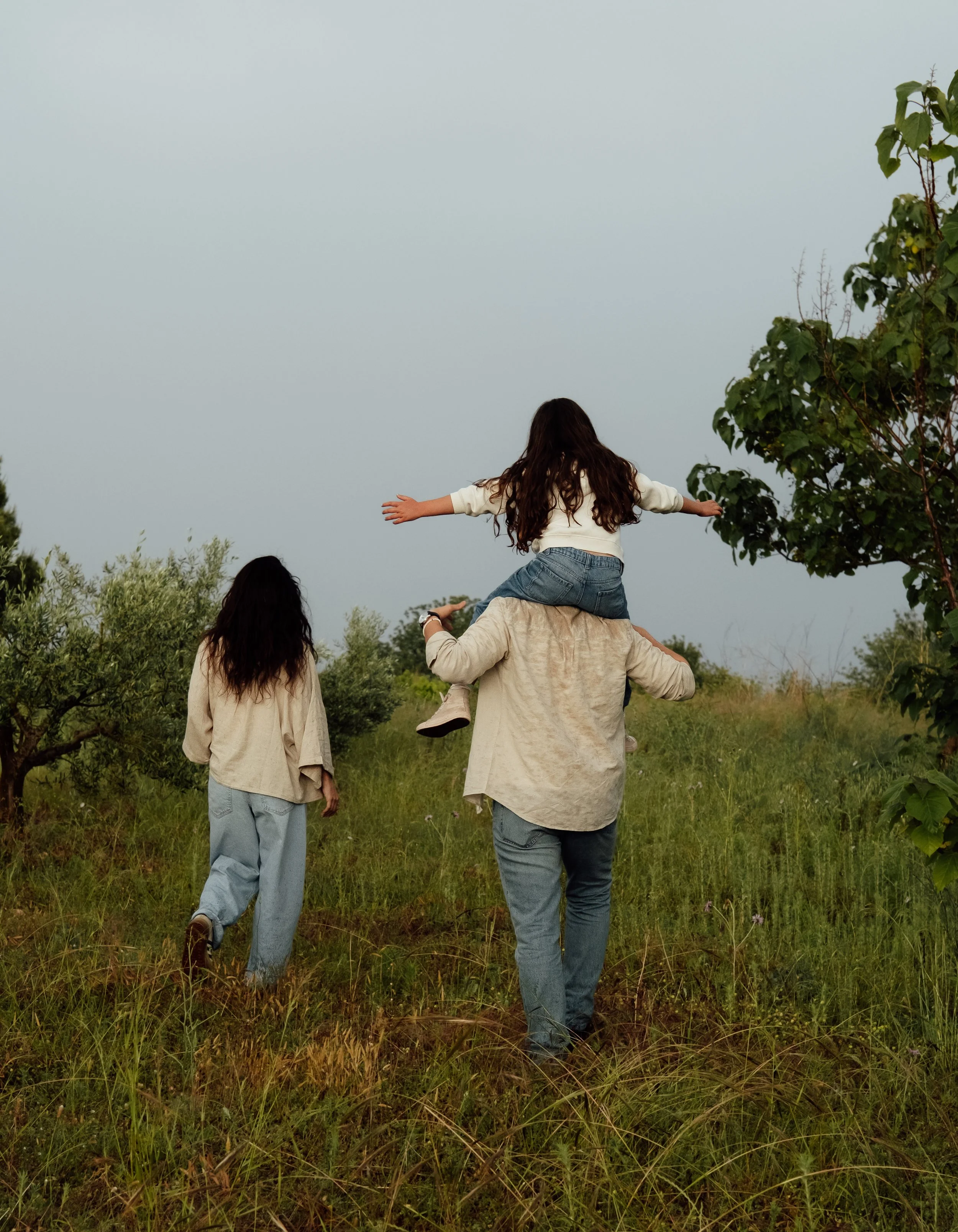 A person carrying a child on their shoulders through a grassy field with trees on either side, under a cloudy sky.