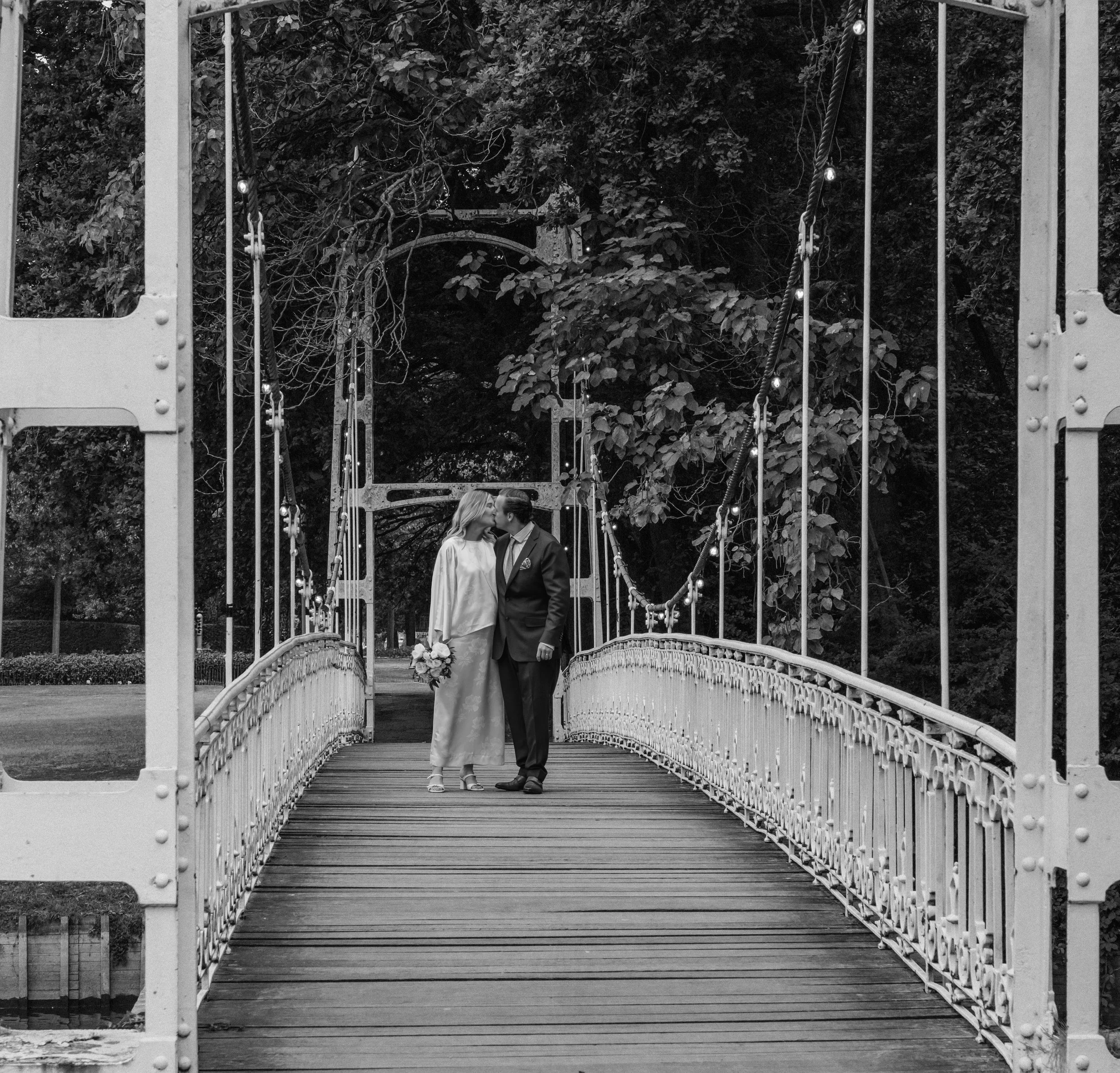 A couple kissing on a bridge surrounded by trees, with string lights hanging above them, in black and white.