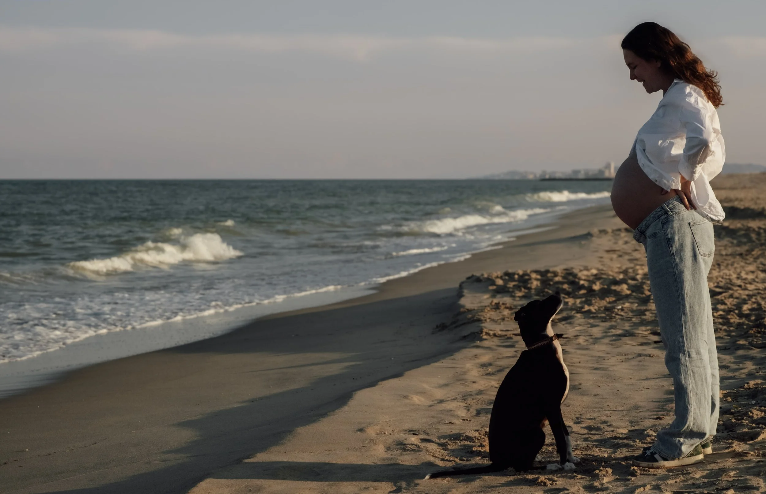 A woman with long brown hair, wearing a white shirt and jeans, stands on a sandy beach with her hands on her pregnant belly, accompanied by a black dog sitting nearby. The ocean waves gently roll onto the shore under a cloudy sky.