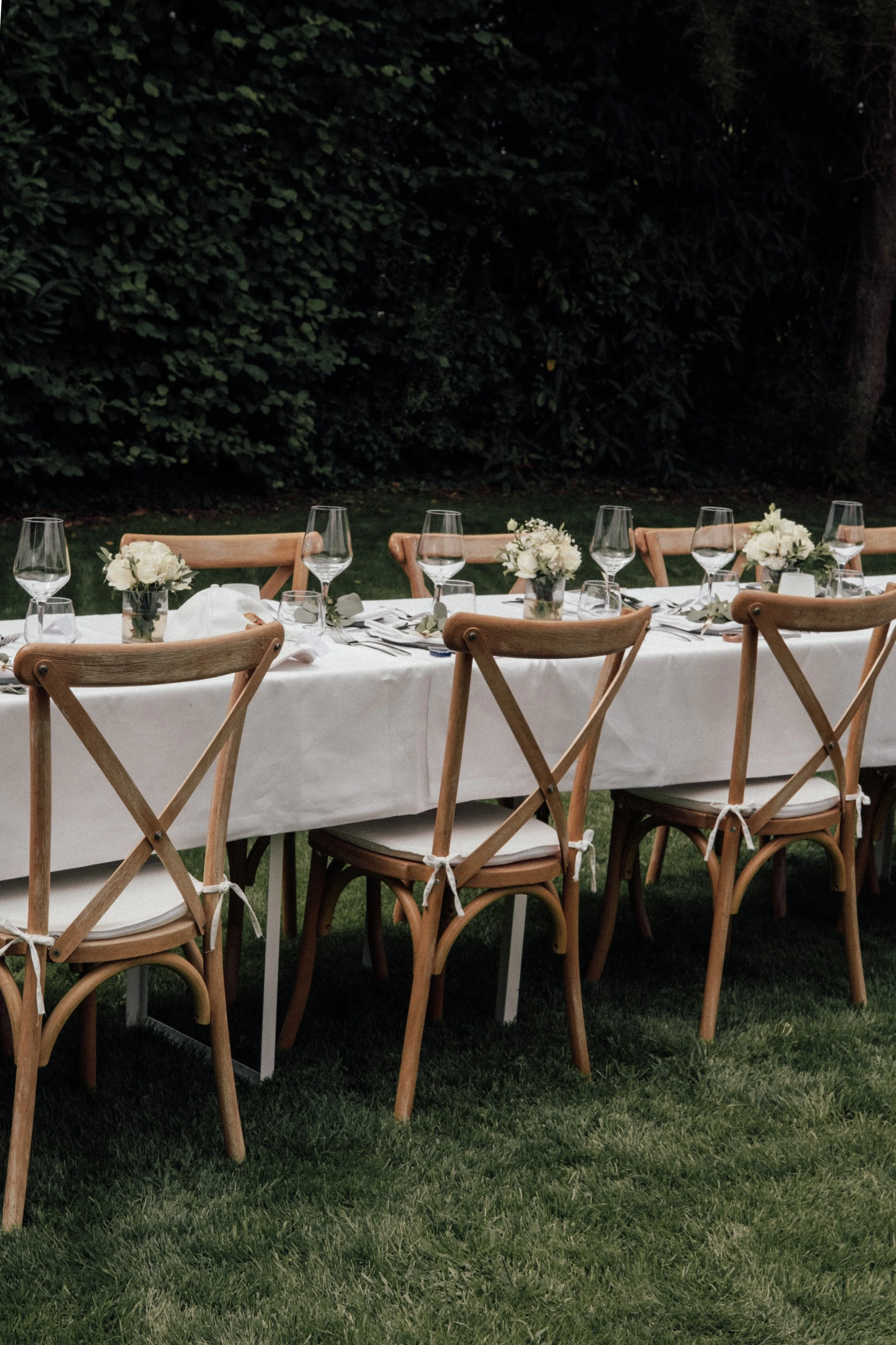 An outdoor dining table set with white tablecloth, floral centerpieces, wine glasses, and napkins. Wooden chairs surround the table, and the scene is in a garden at dusk with dark green foliage in the background.