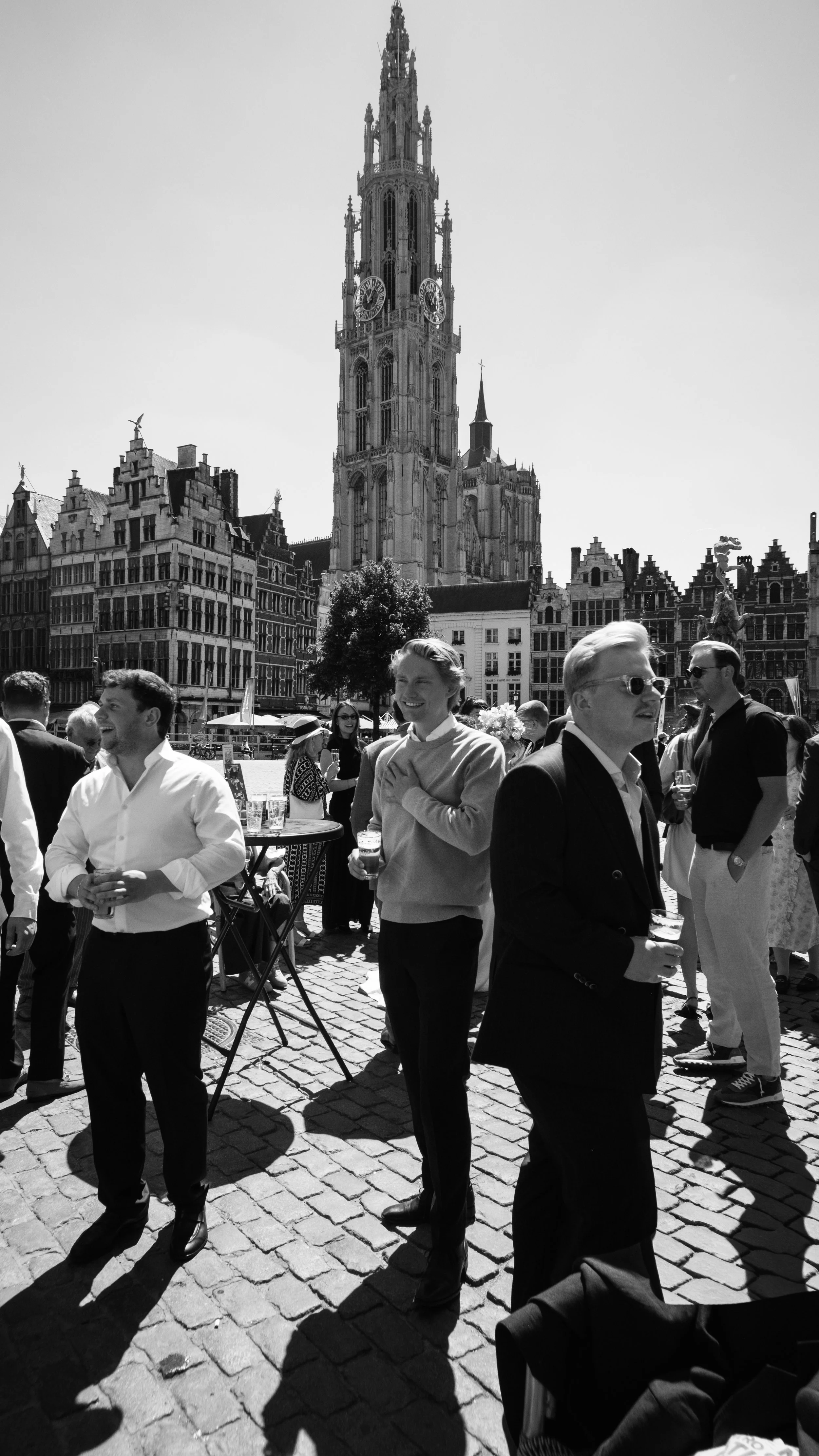 People standing and socializing in a lively outdoor square with historic buildings and a tall clock tower in the background.