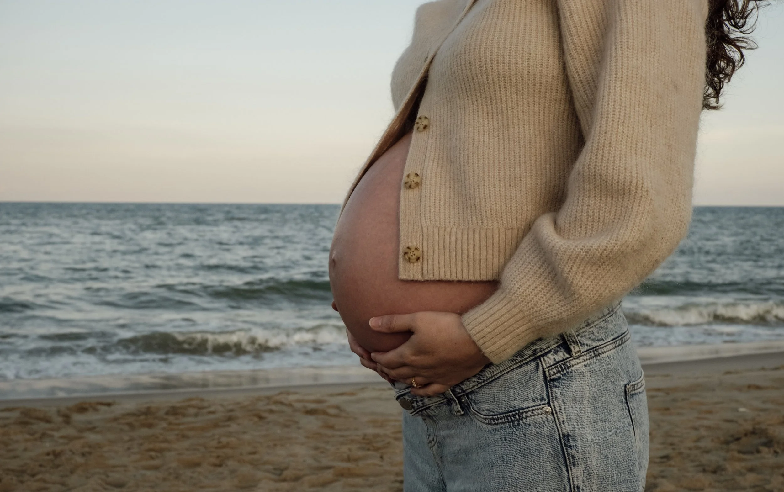 A pregnant woman in a beige sweater and jeans standing on the beach holding her belly with both hands, with the ocean and sky in the background.