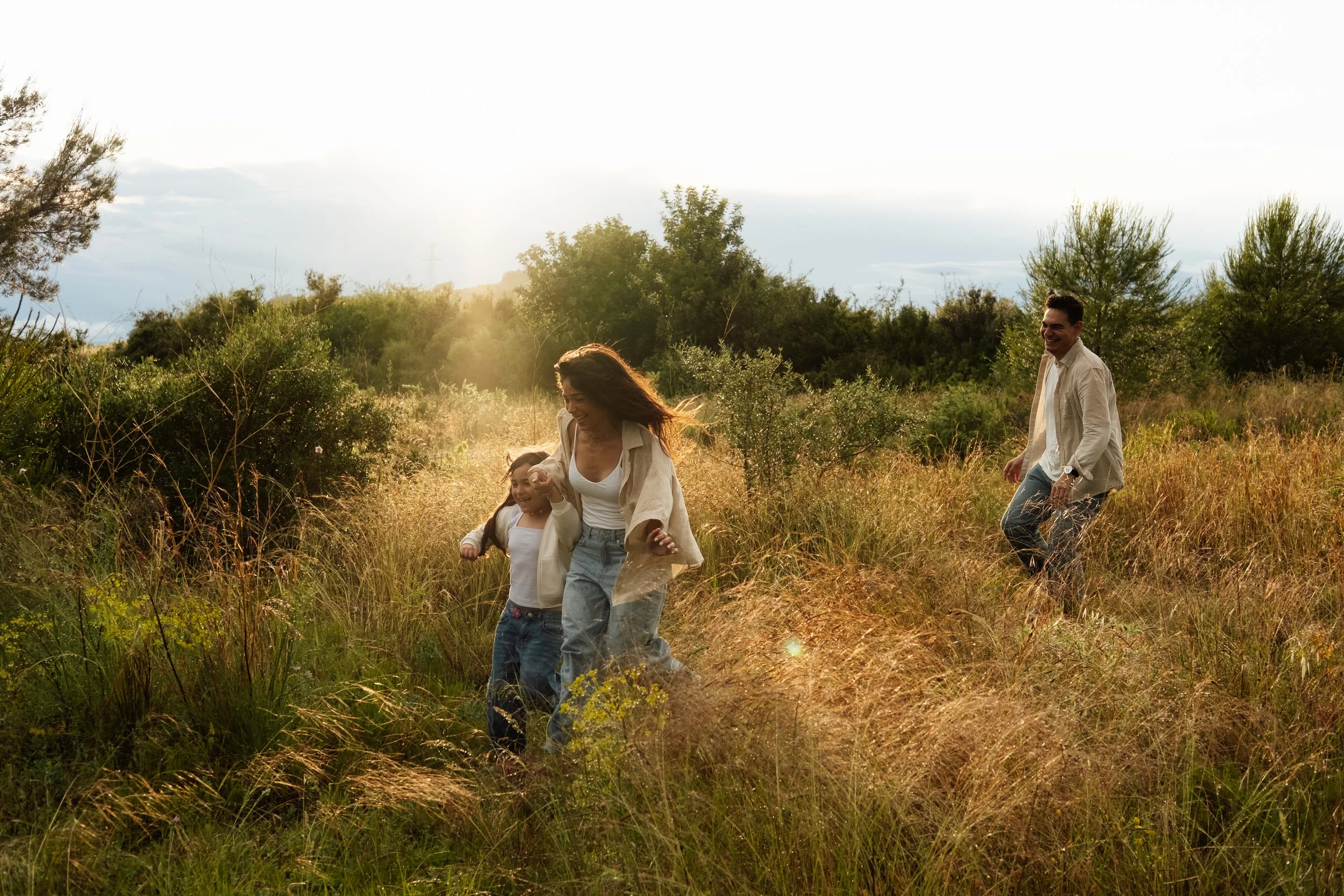 A family of four, including two children, walking and playing in a sunlit grassy field surrounded by trees, enjoying outdoor and nature.