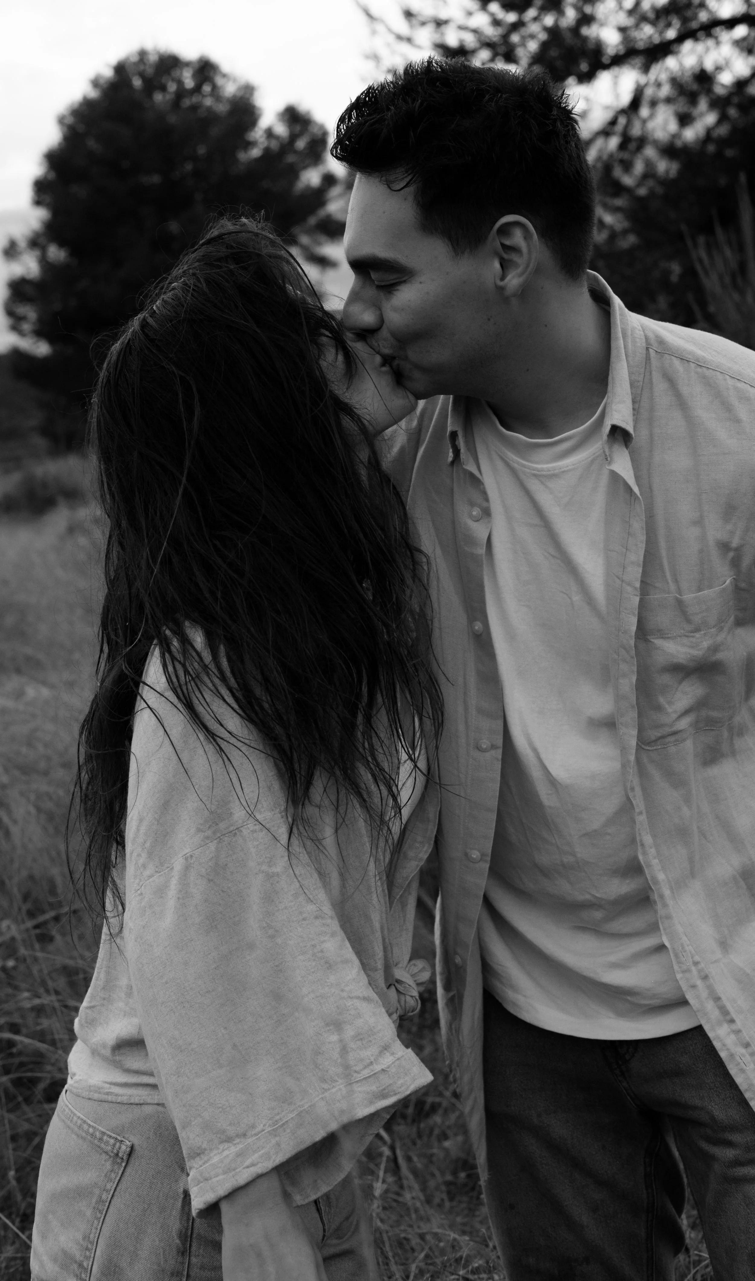A black and white photo of a couple kissing outdoors, with trees and grass in the background.