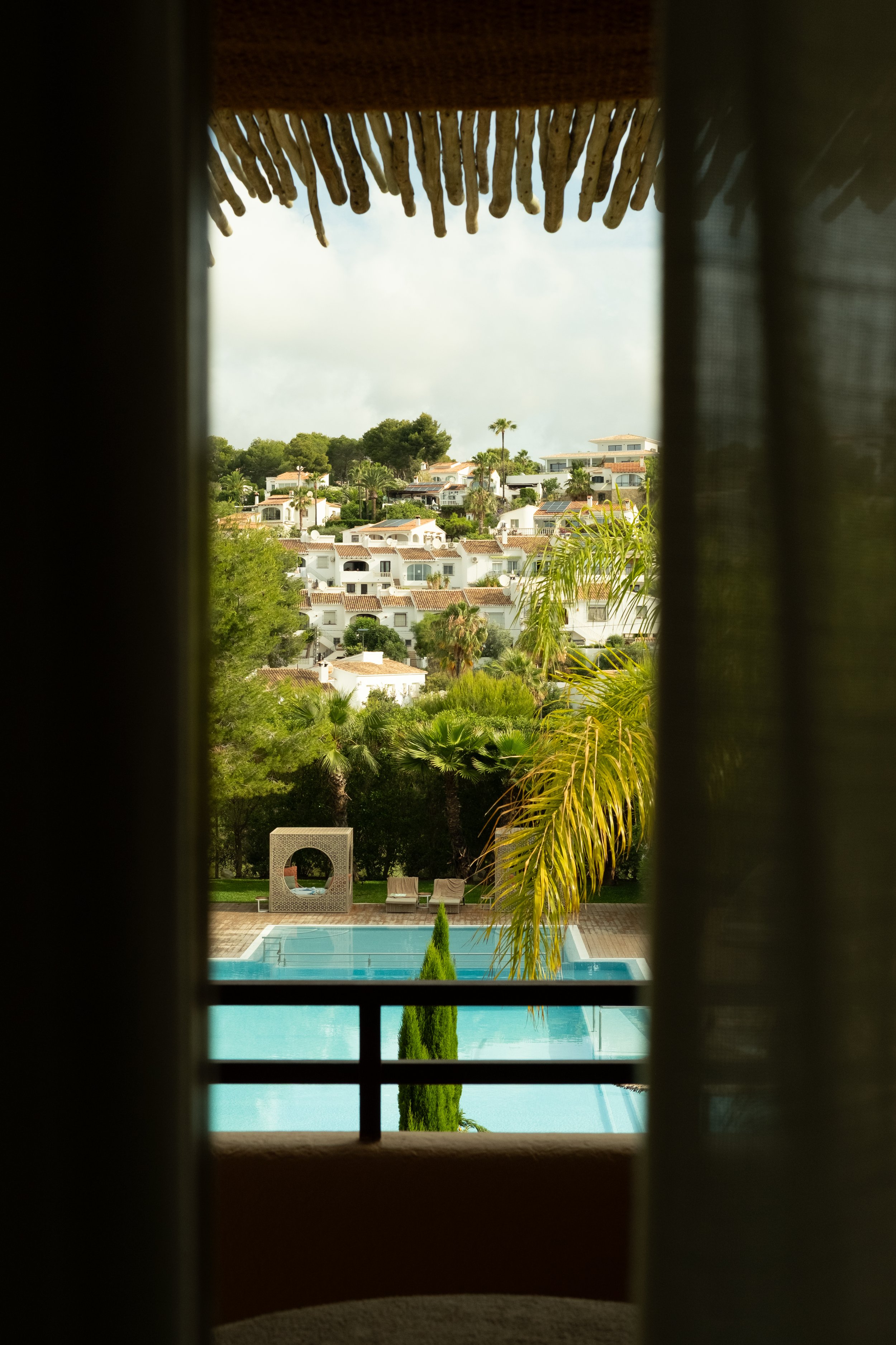 View of a swimming pool surrounded by trees and chairs, seen through a window with curtains at the top and bars at the bottom.