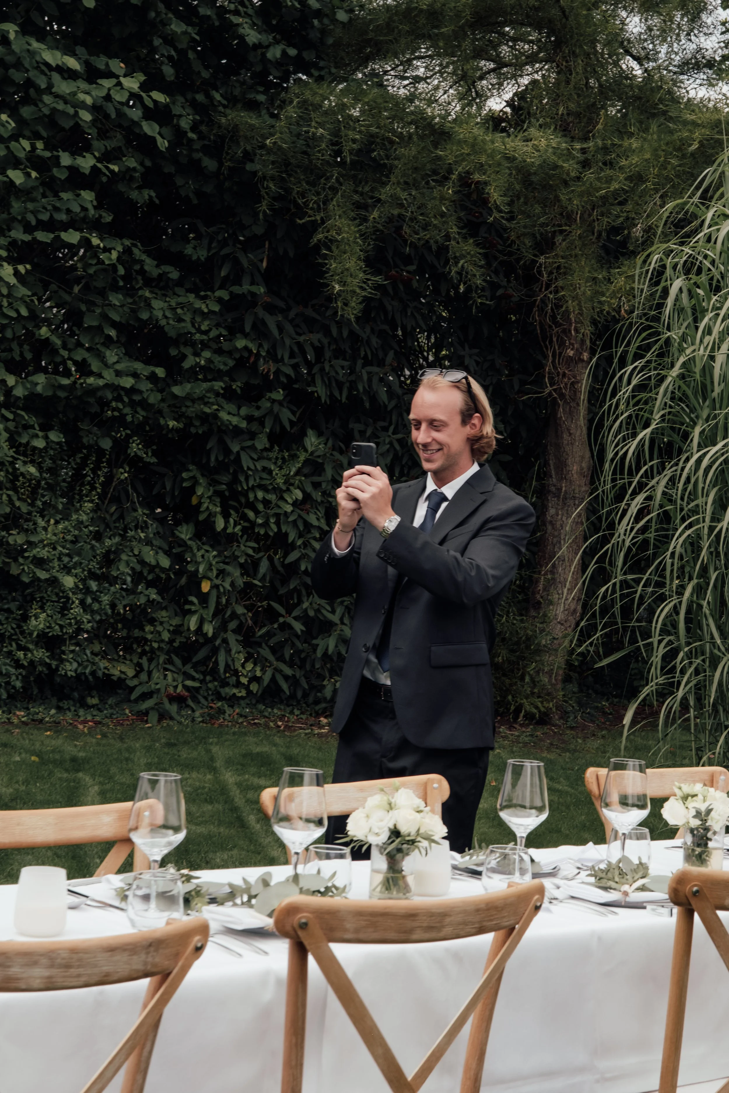 A man in a black suit and tie, with sunglasses on his head, is smiling and looking at his phone outdoors in a garden setting, with a table set for a meal in the foreground.