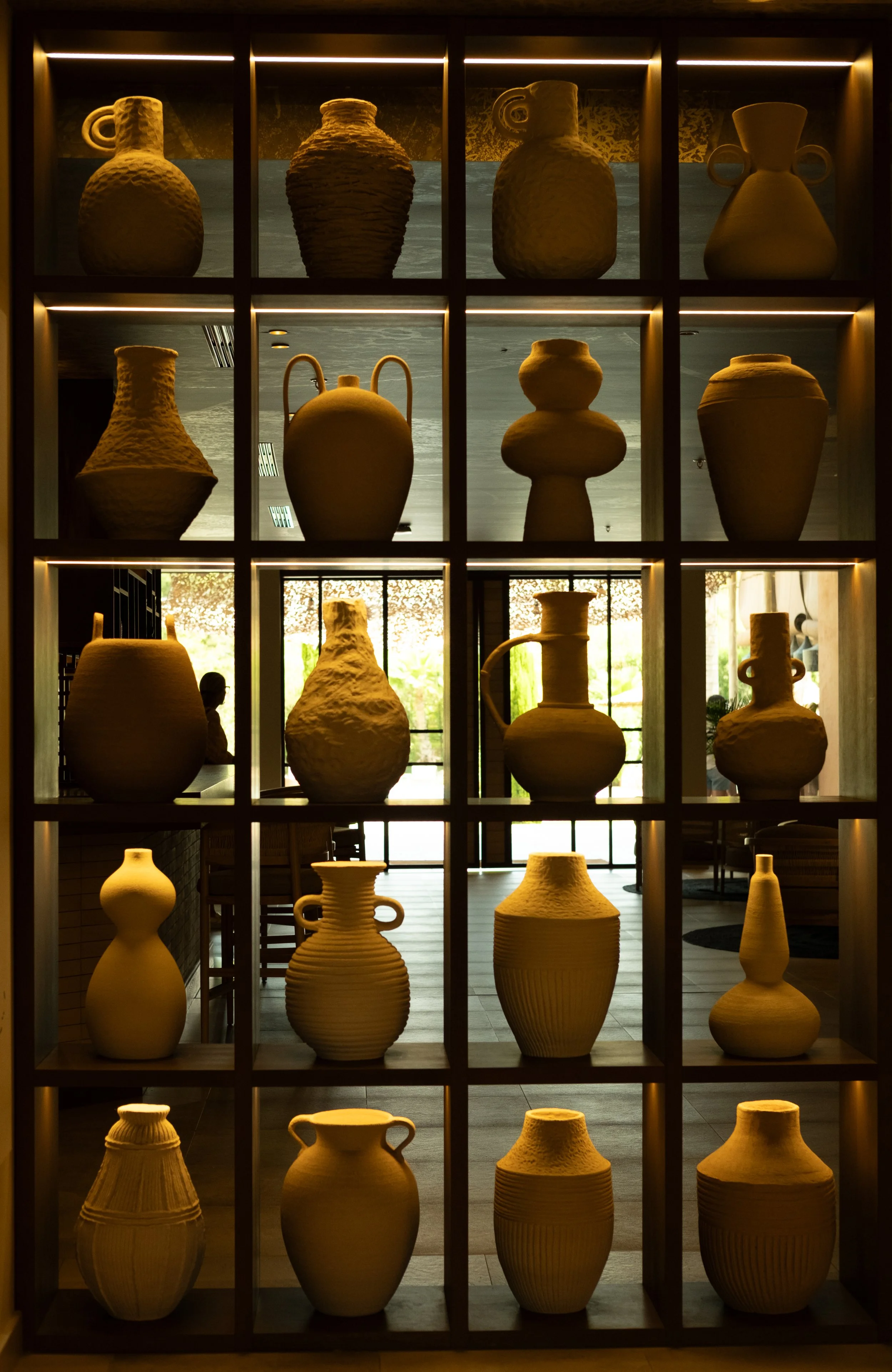 Display of various ceramic vases on wooden shelves inside a building with large windows, showing outdoor greenery.