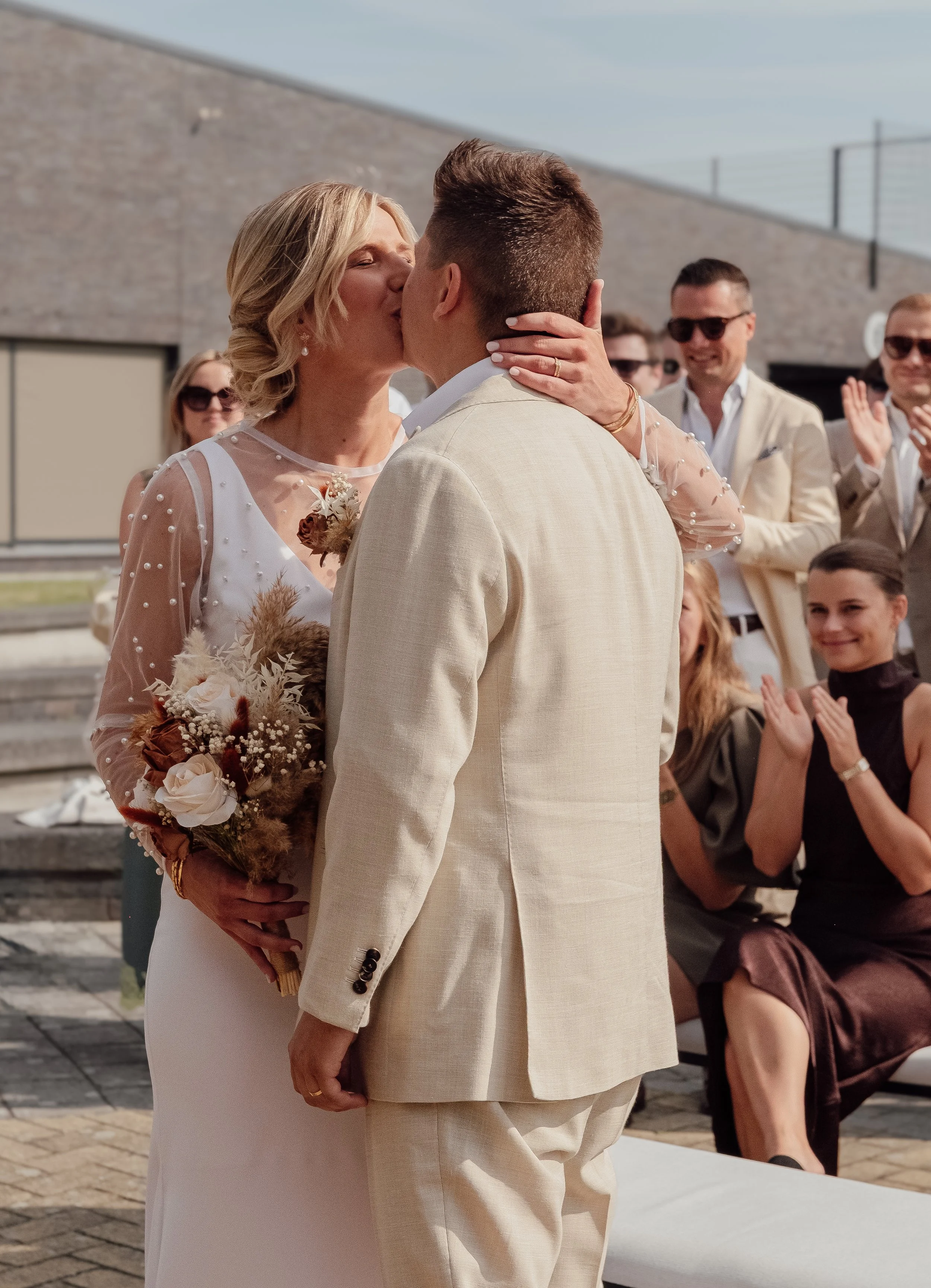 A couple is kissing, celebrating their wedding outdoors with friends watching and applauding.