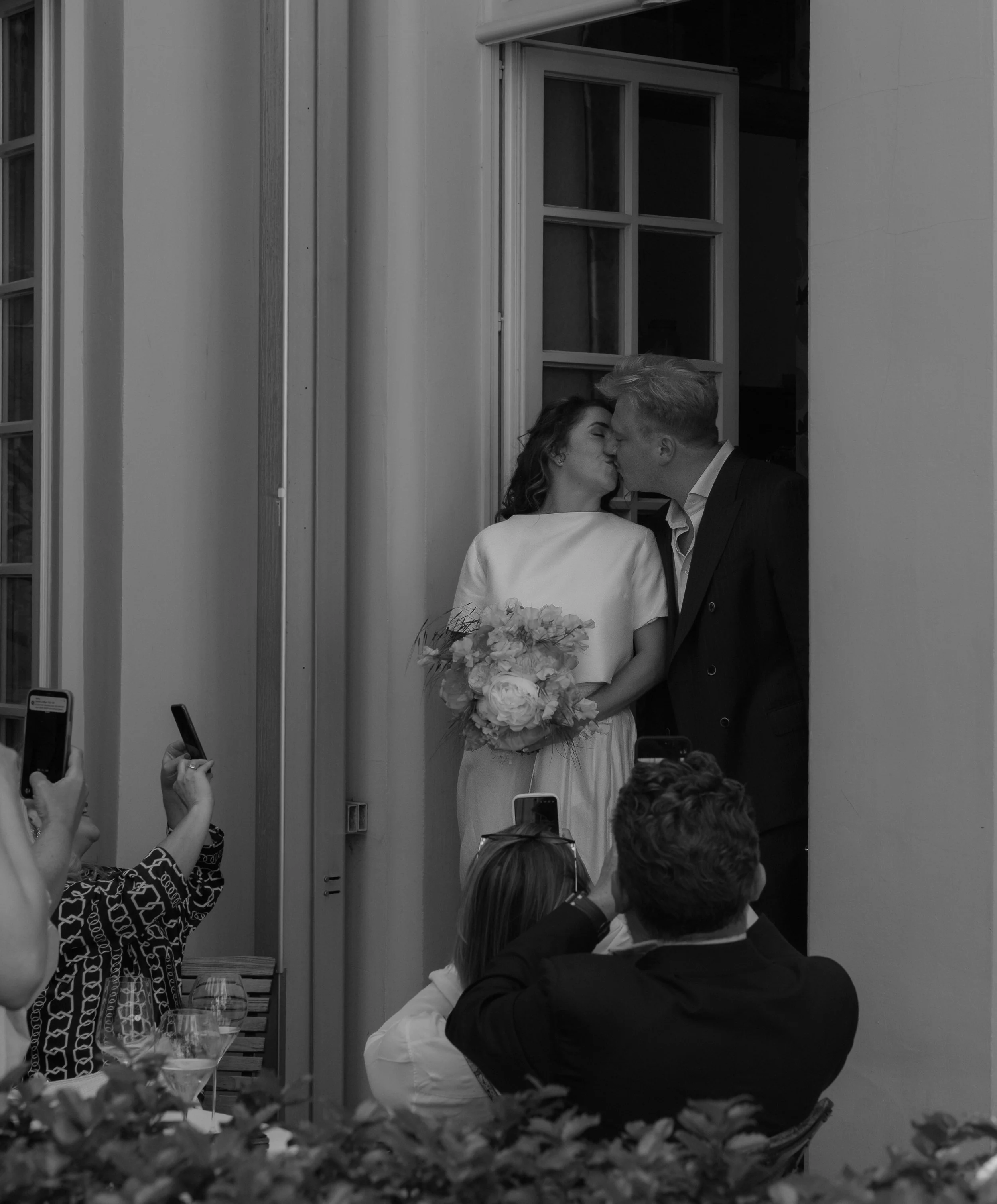 A black-and-white photograph of a newlywed couple sharing a kiss at a wedding reception, surrounded by guests taking photos and holding drinks. The bride holds a bouquet of flowers, and the groom is dressed in a dark suit. They stand near an open win