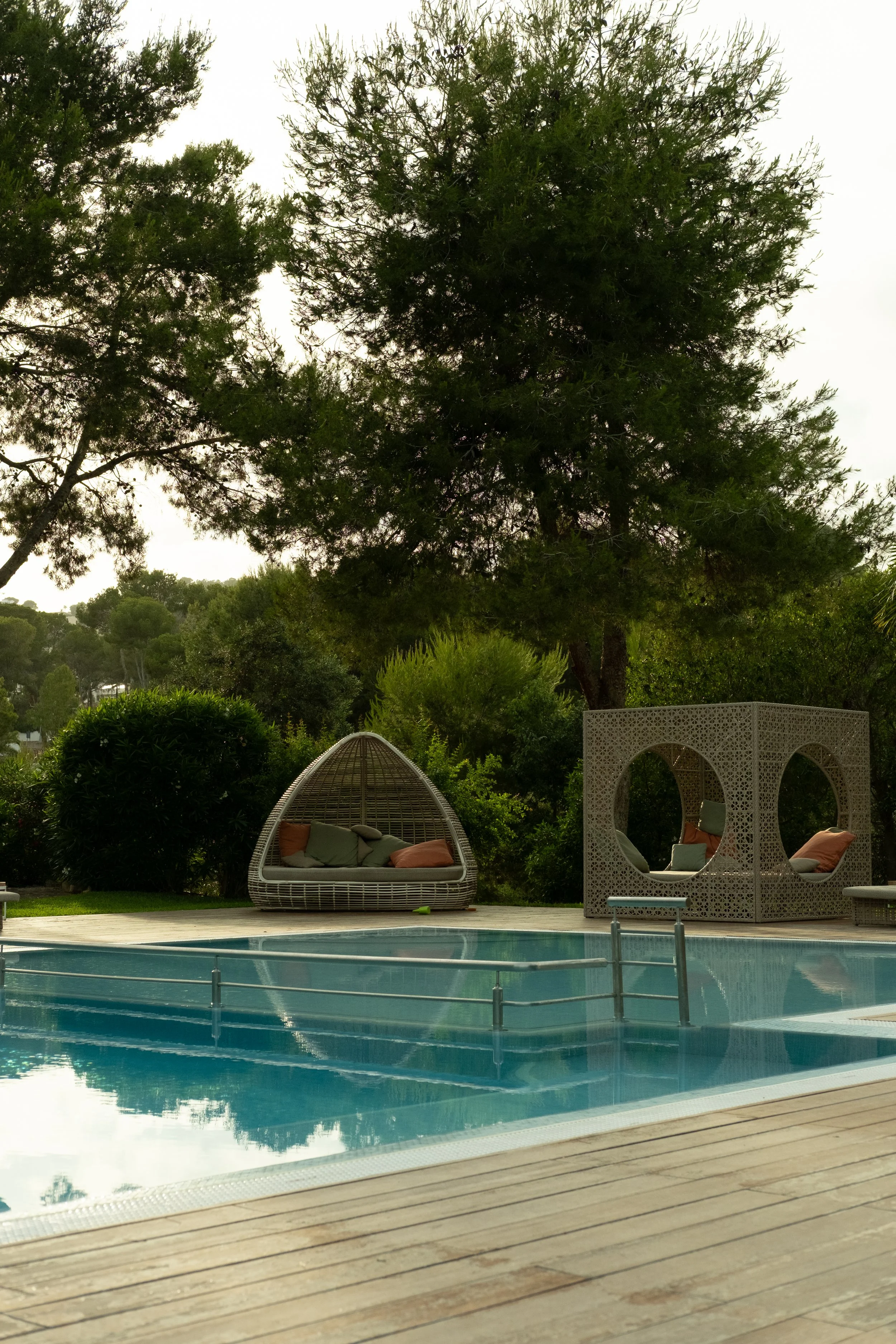 A backyard scene with a swimming pool, outdoor wicker lounge chair with pillows, decorative white lattice structure with circular cutouts, lush green trees, and cloudy sky in the background.