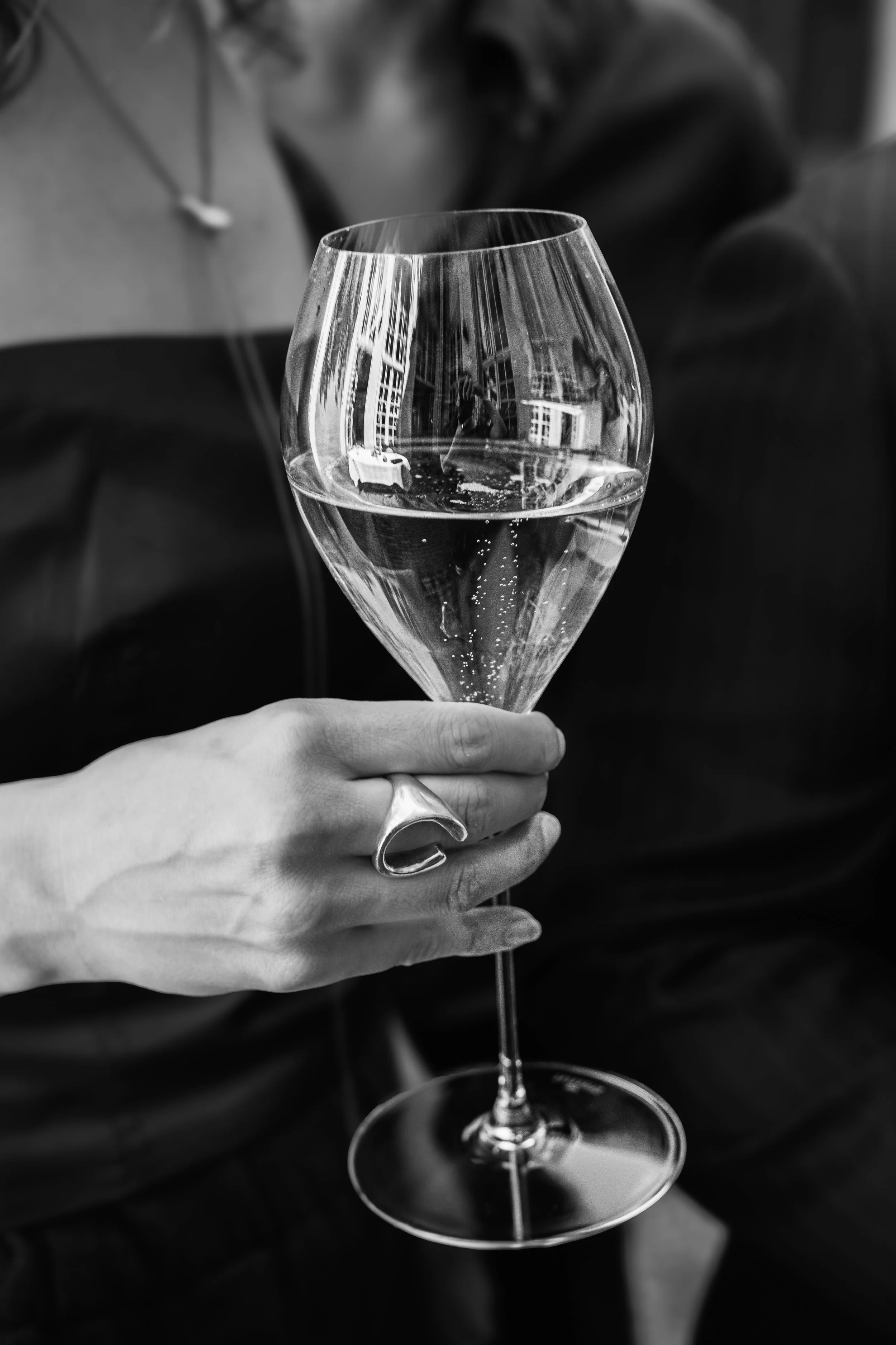 A woman holding a champagne flute filled with sparkling wine, with a reflection of a building visible in the glass, in black and white.