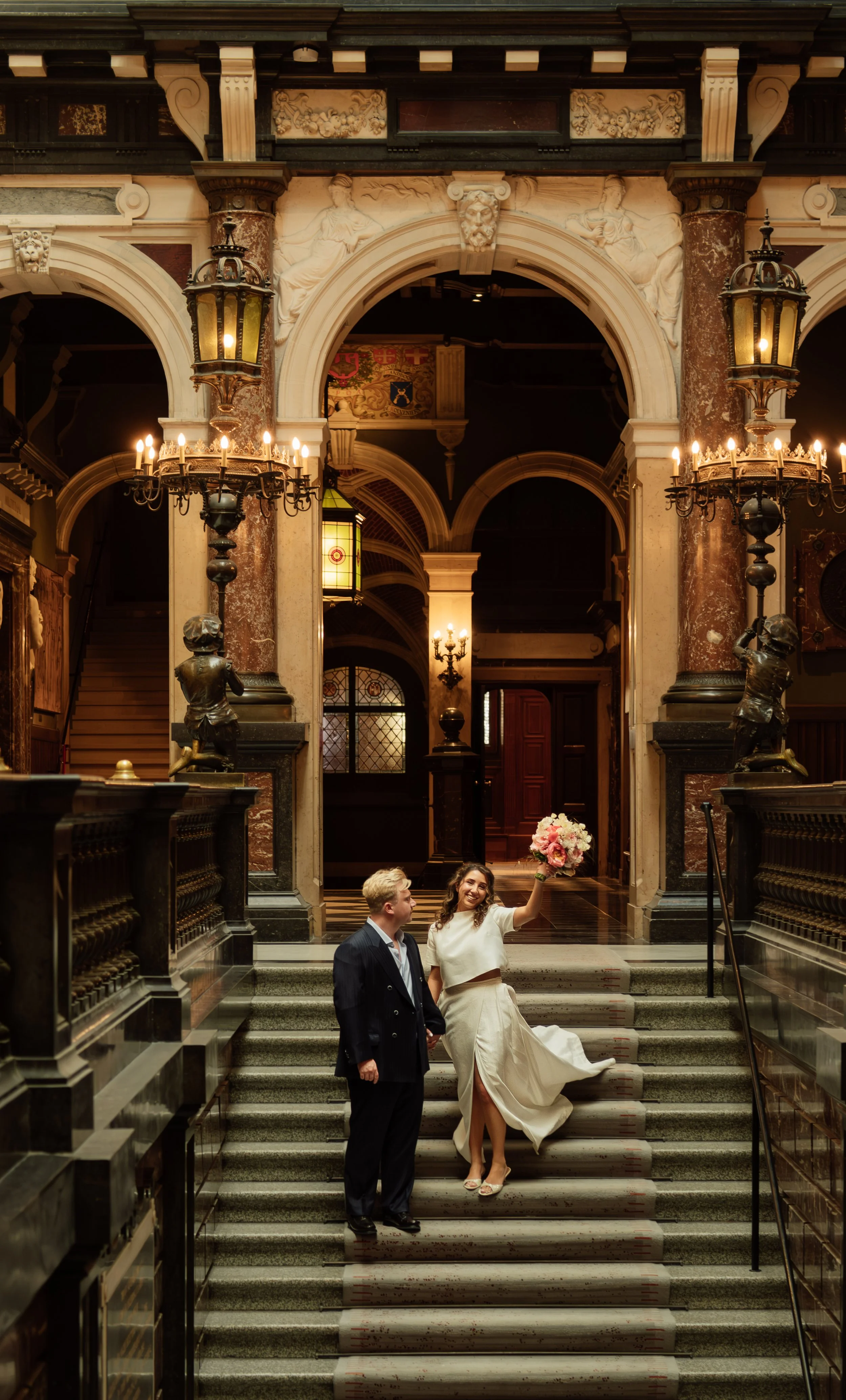 A wedding scene inside an elaborate, historic building with marble columns, ornate sculptures, and chandeliers. A bride in a white gown holding a bouquet smiles and waves, standing on stairs next to a groom in a dark suit. The setting features intric