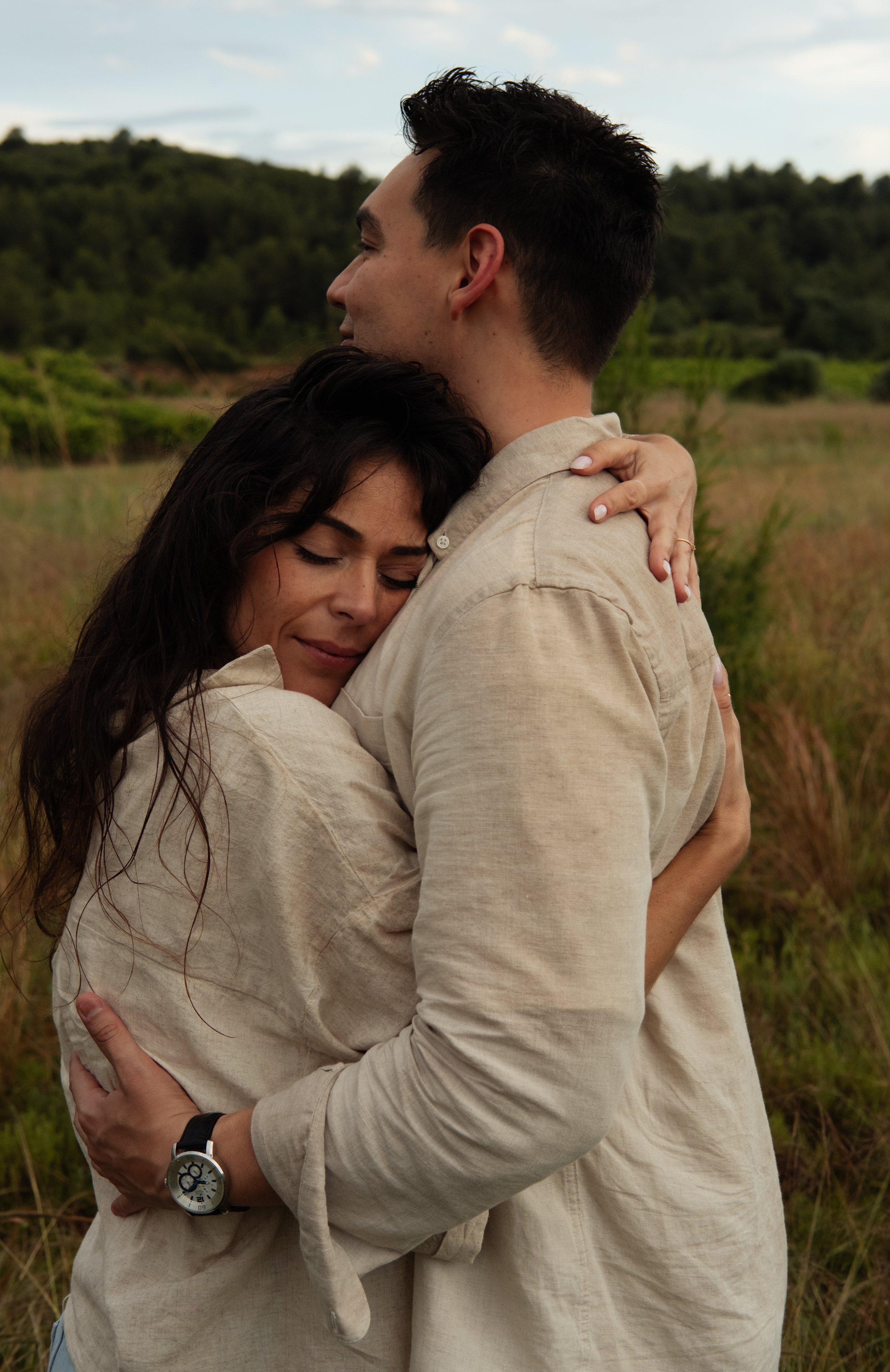 A man and woman hugging outdoors in a grassy field with trees and a cloudy sky in the background.