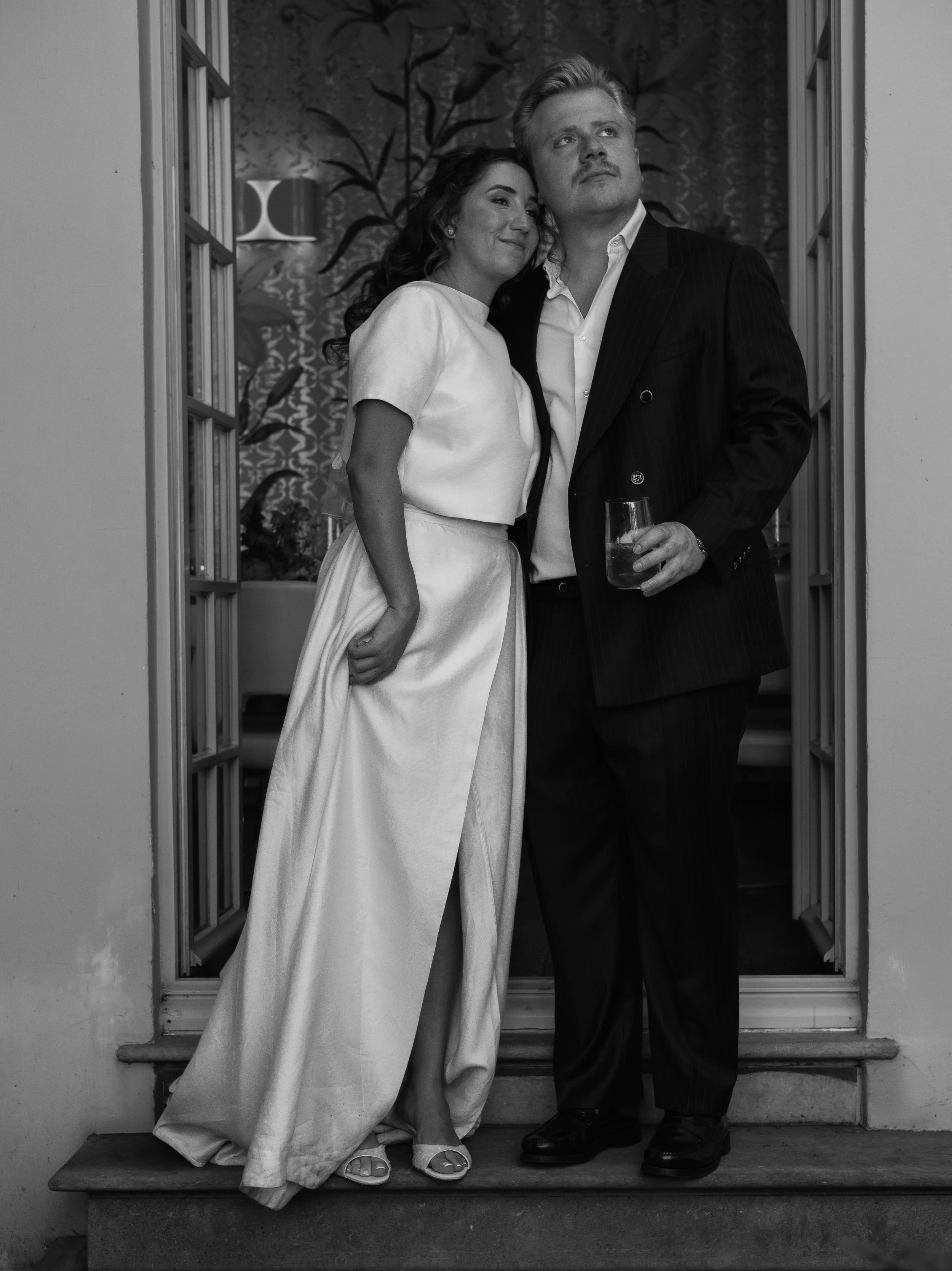 A black and white photo of a couple standing on a front porch. The woman is leaning her head on the man's shoulder, both are smiling, and the man is holding a glass of drink. The woman is in a light-colored dress, and the man is in a dark suit. There
