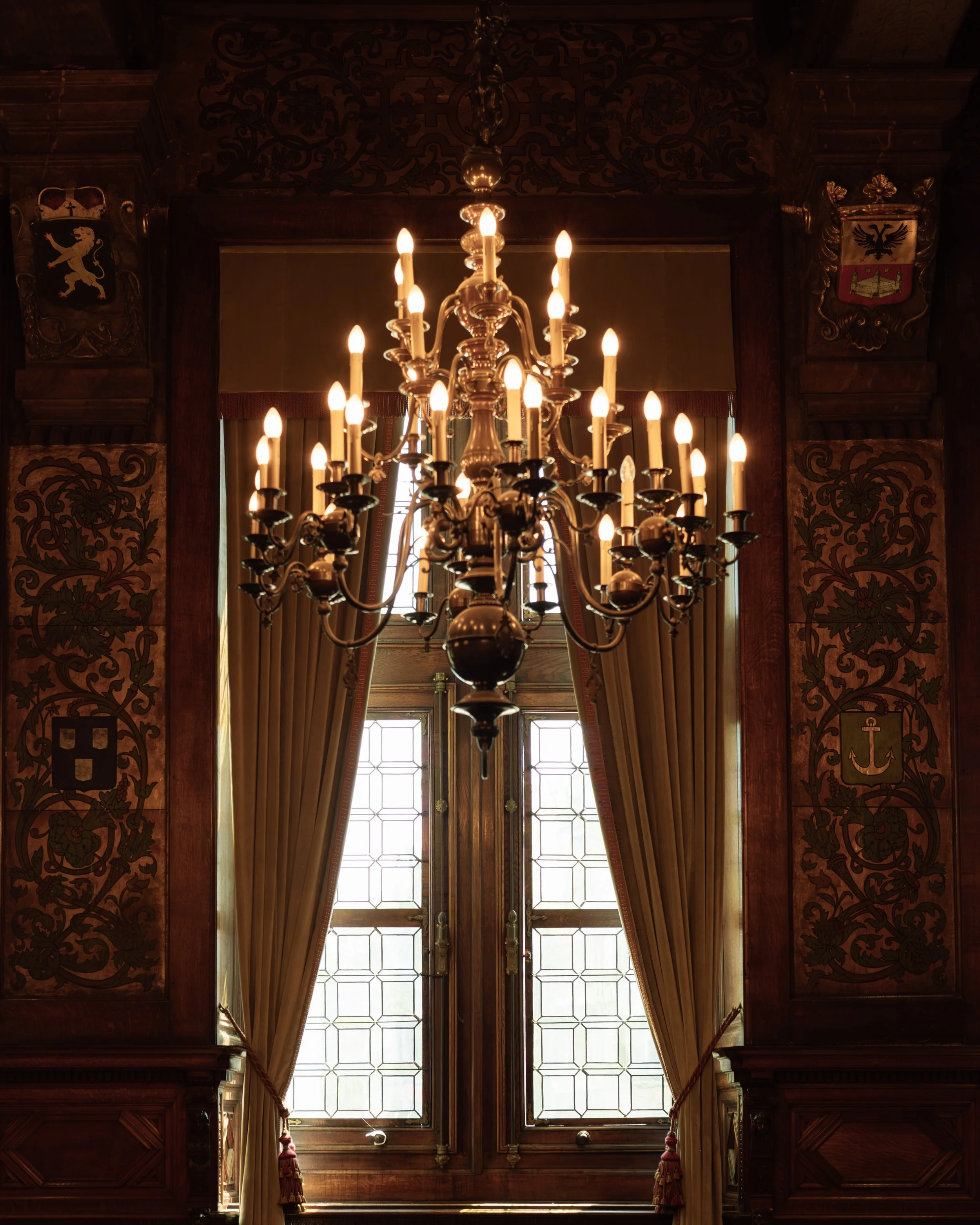 Elegant room with a large, ornate chandelier hanging from the ceiling, positioned in front of tall windows with curtains. Wooden paneling and crests with heraldic symbols adorn the walls.