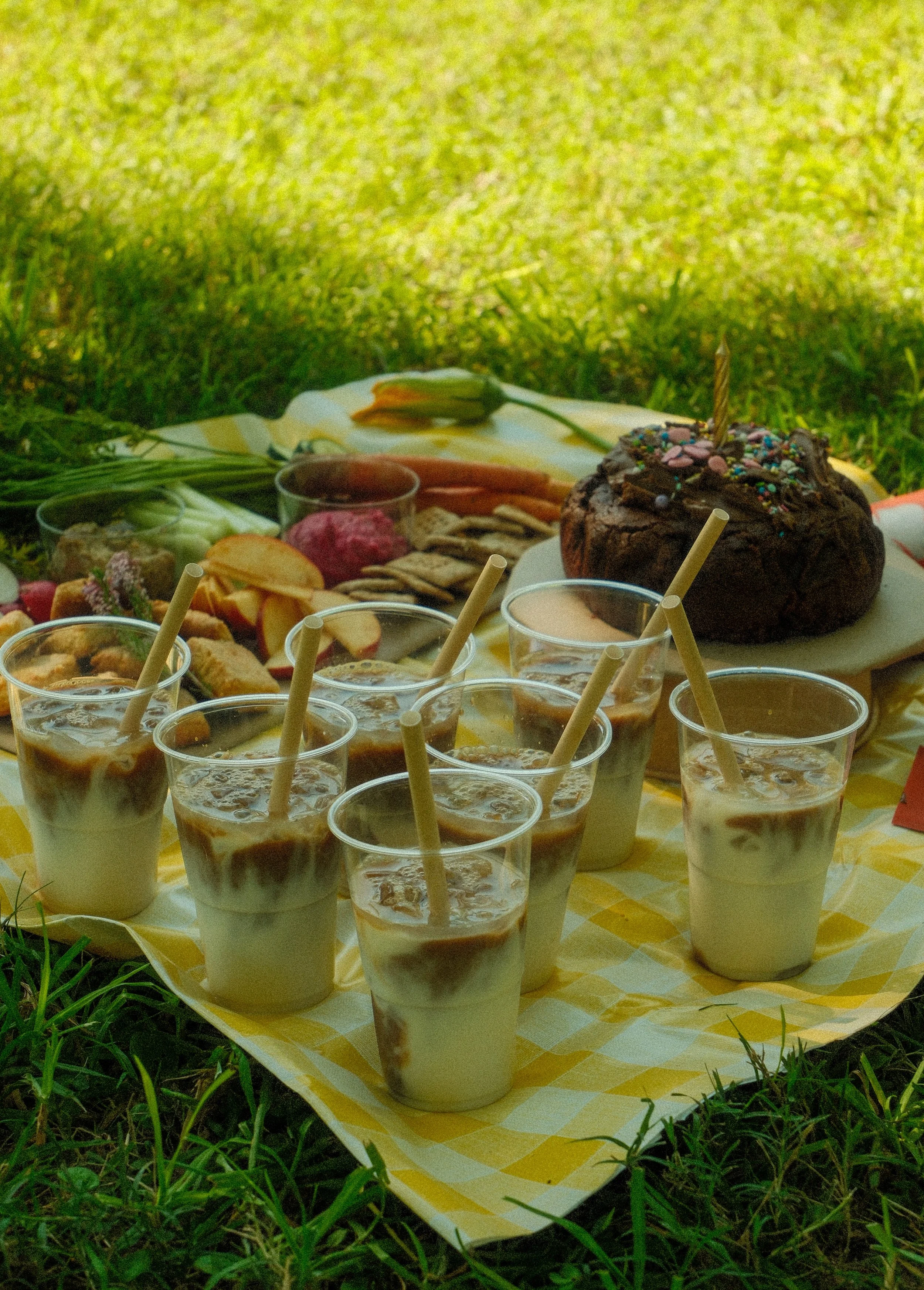 A picnic table set on grass with a checkered yellow and white tablecloth, topped with a chocolate cake with a birthday candle and rainbow sprinkles, eight glasses of iced coffee or milkshakes with straws, and various snacks including fruit, cookies, 