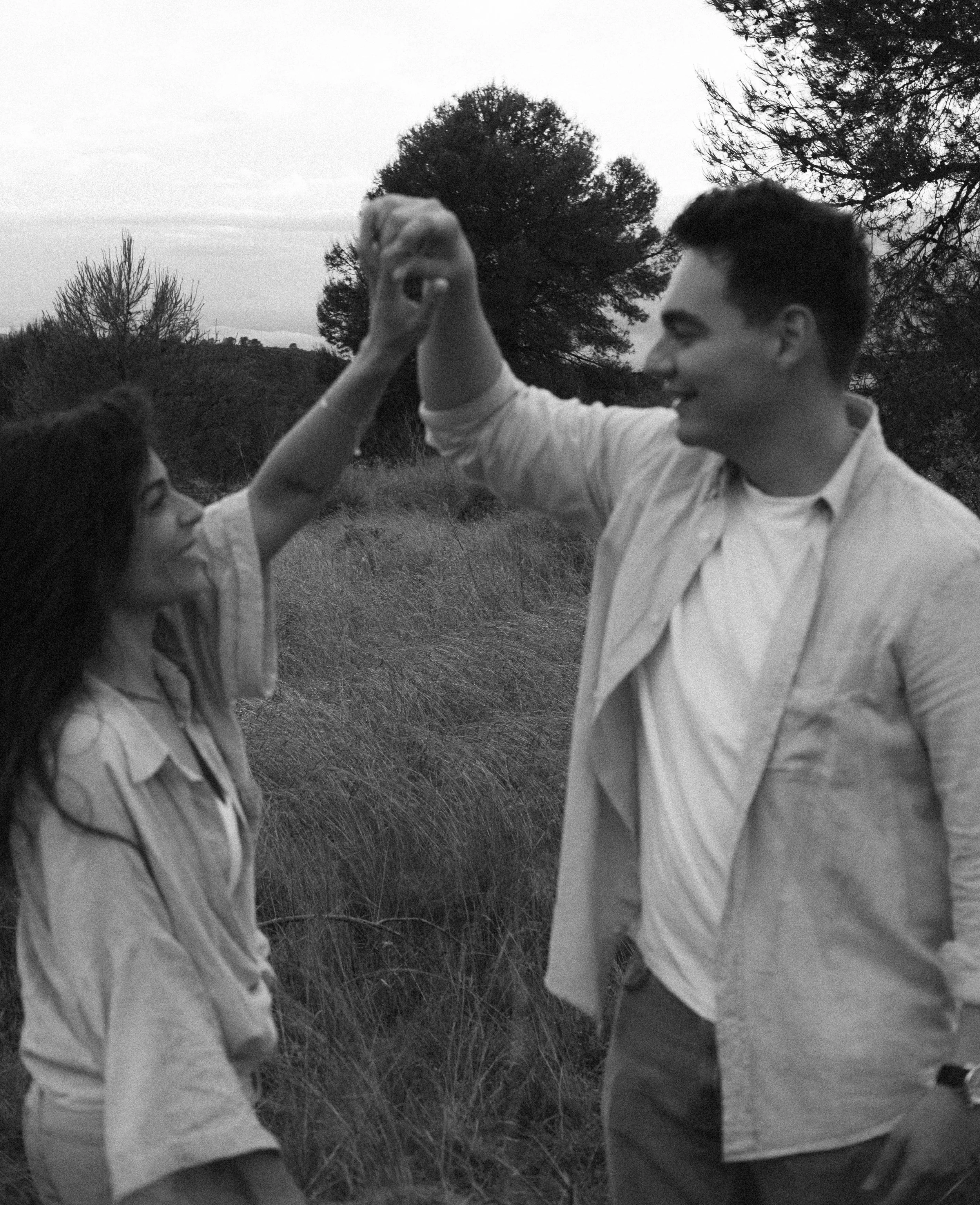 A black and white photo of a young man and woman holding hands in an outdoor field with trees in the background.