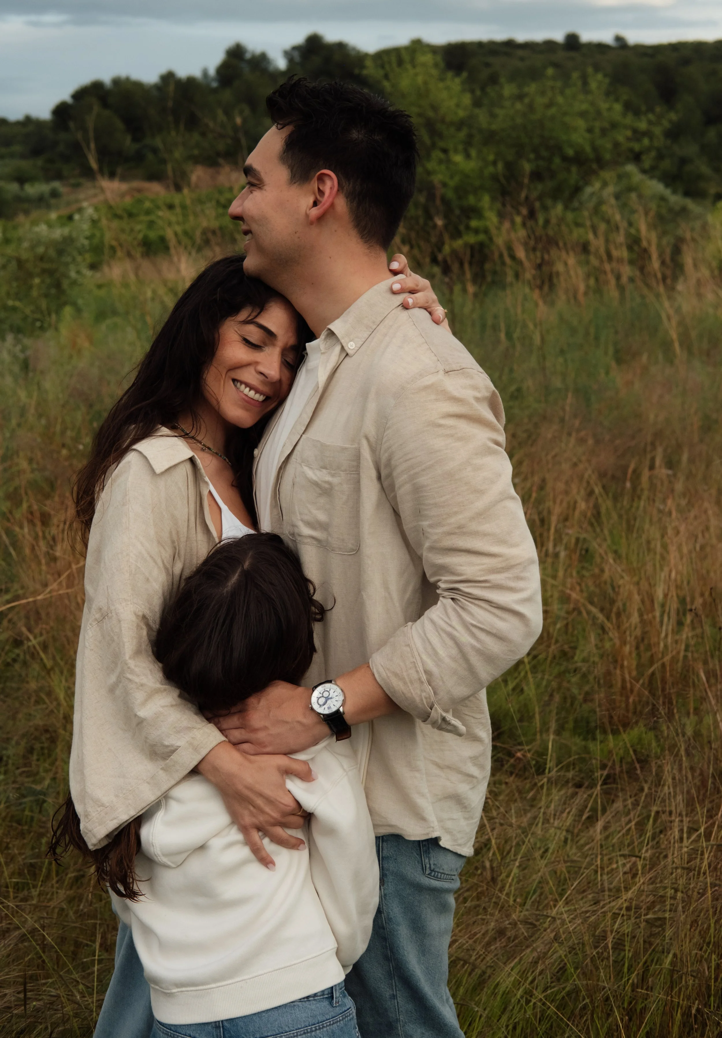 A happy couple sharing a warm embrace outdoors in a grassy field, with lush green trees and a cloudy sky in the background.