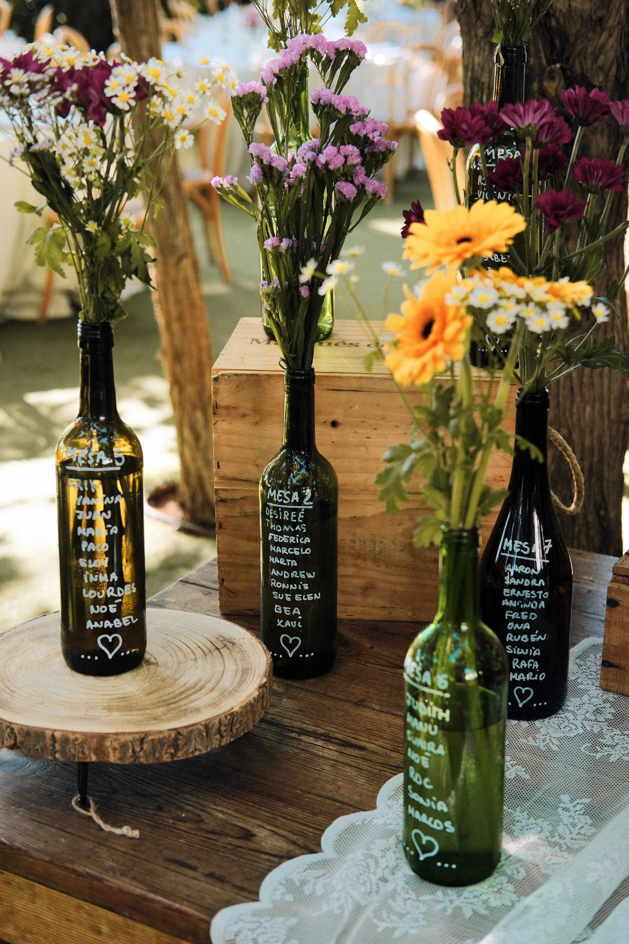Vases made from green and dark glass bottles with handwritten lists of names on them, arranged with colorful flowers on a wooden table. The background is softly blurred with a natural outdoor setting.