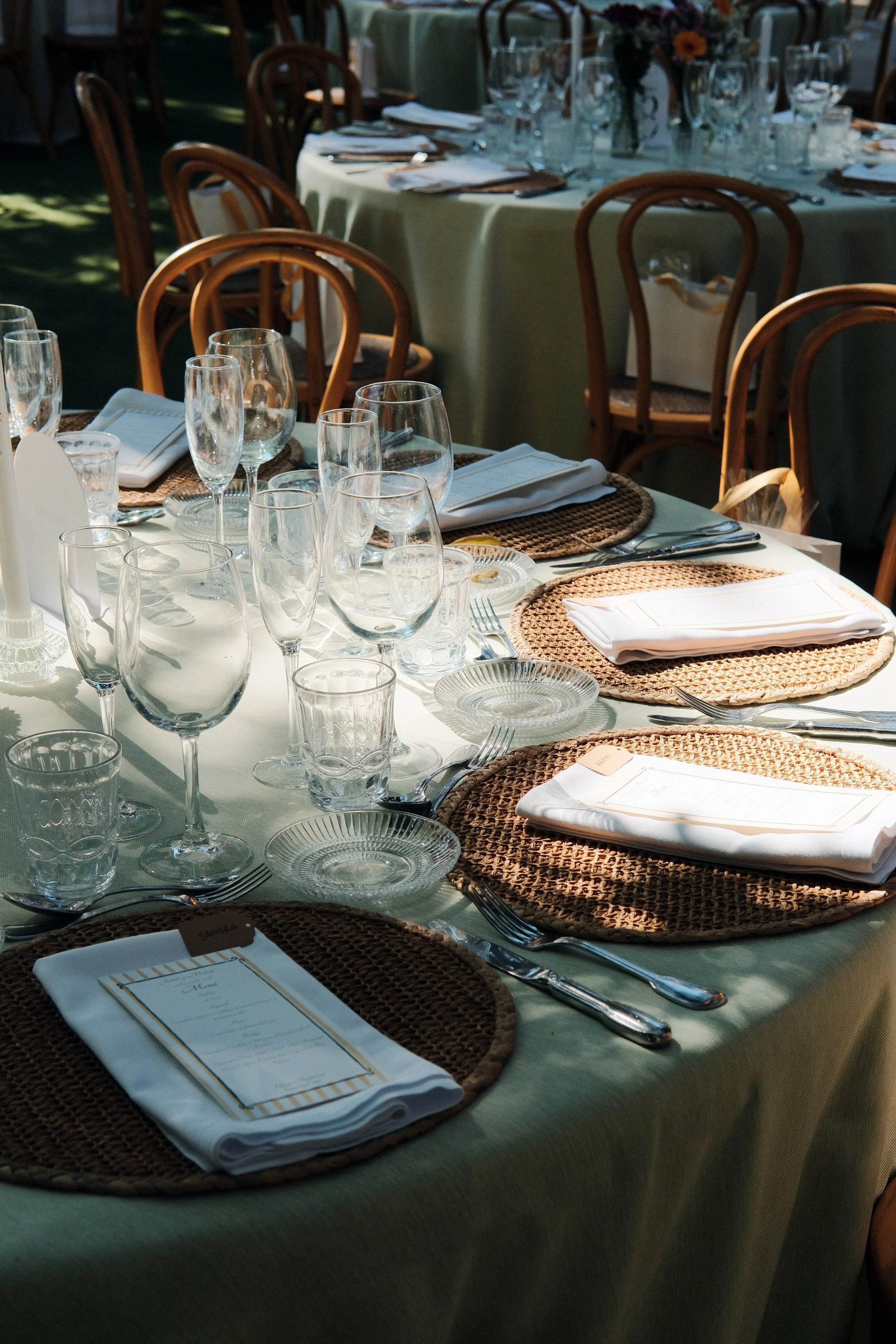 Set dining table with white tablecloth, woven placemats, neatly folded white napkins, clear glassware, silverware, and menus, arranged for an outdoor or indoor event.