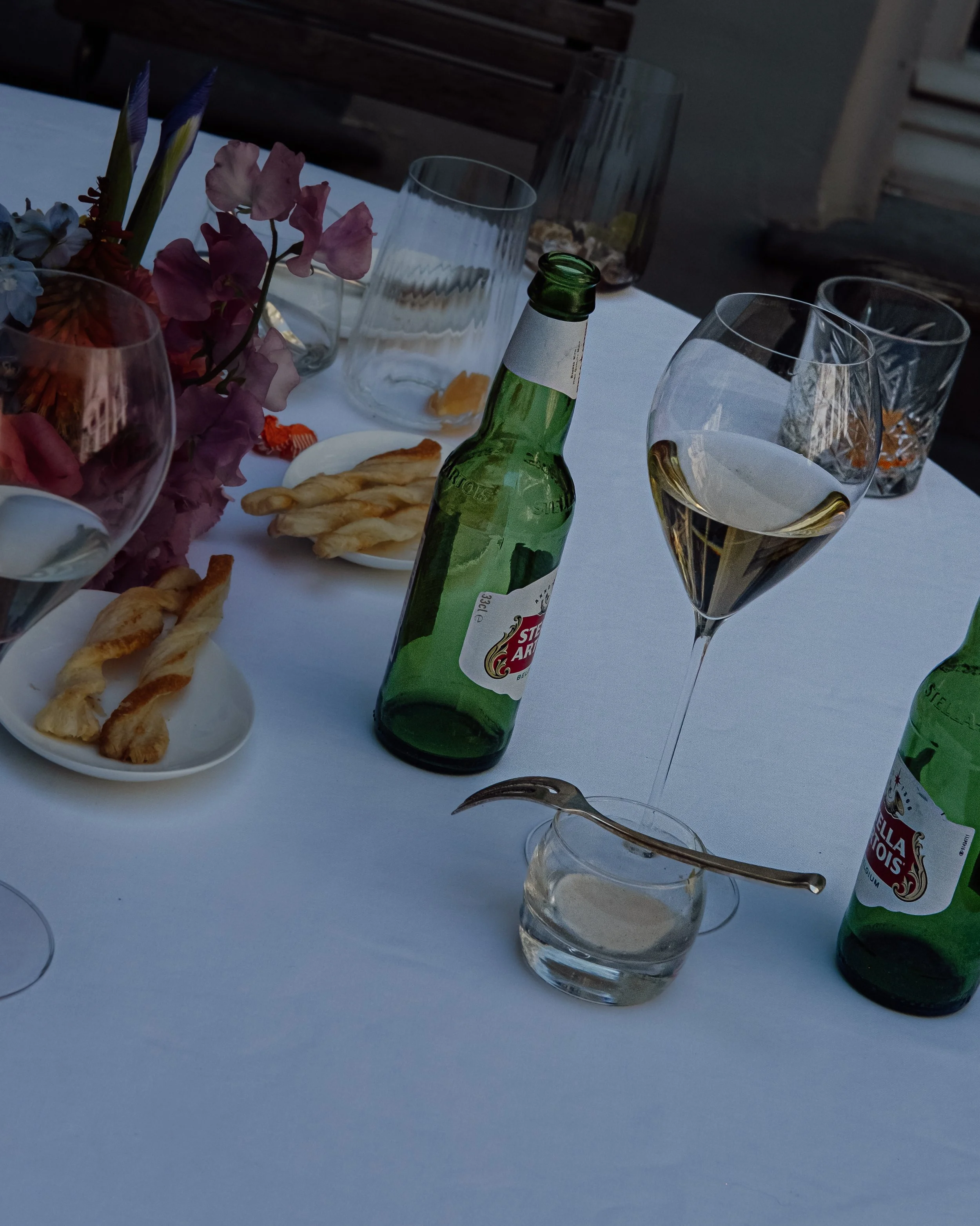 Table set with flowers, several glasses, a green glass bottle, and crispy breadsticks at an outdoor gathering.