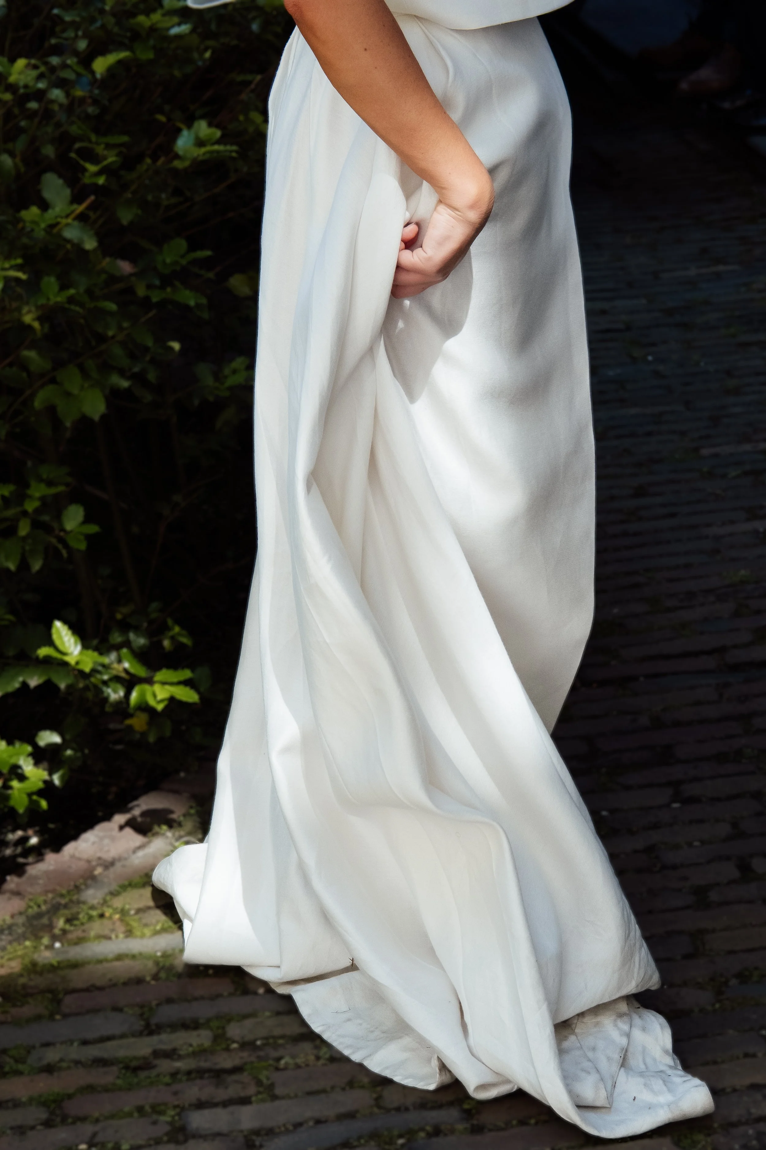 Close-up of a person wearing a long, flowing white dress, standing outdoors on a cobblestone path with green bushes in the background.