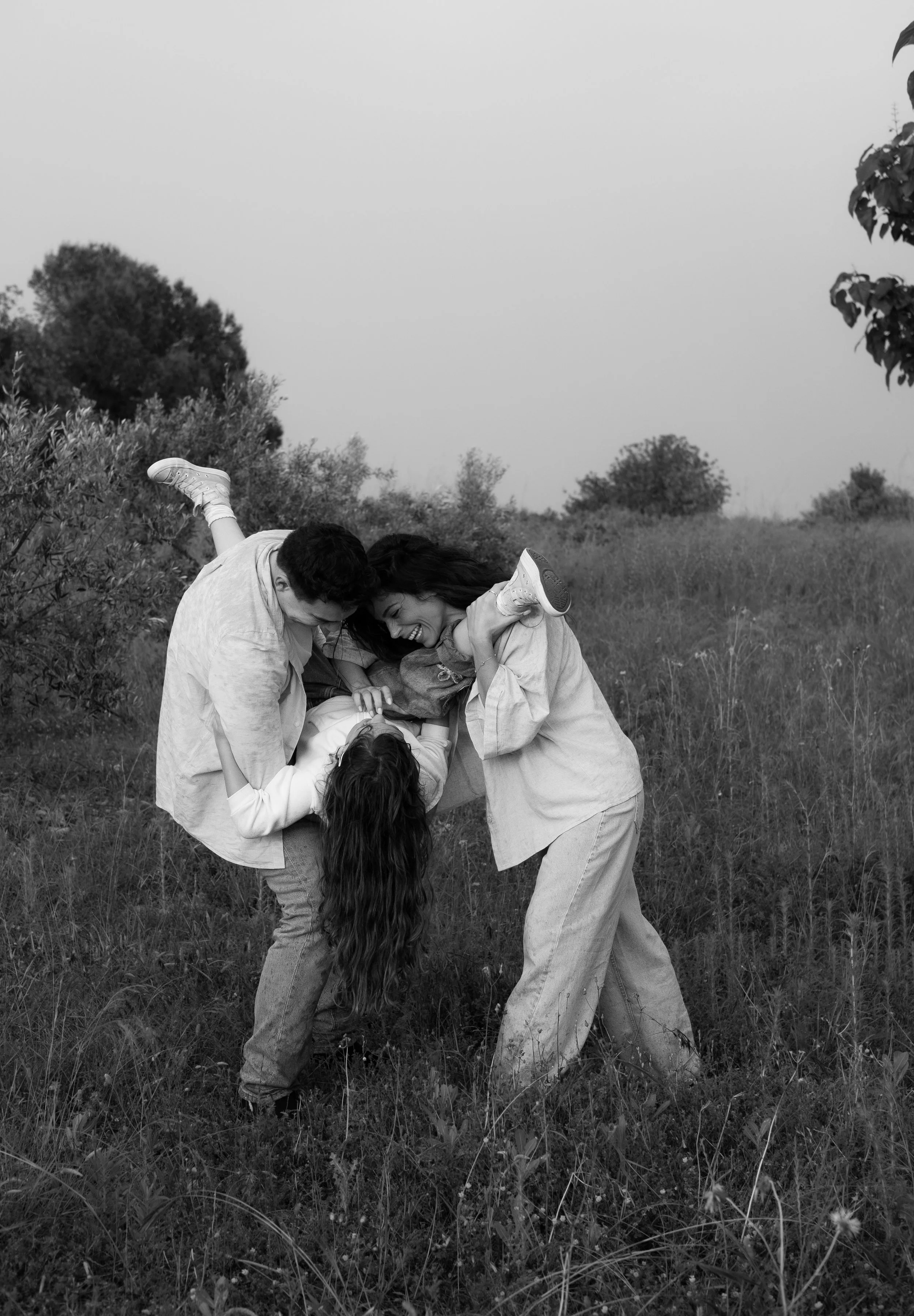 Two people are outdoors in a grassy field, engaging in a playful, maypole dance. They are laughing and holding a long piece of fabric or ribbon, with a child hanging upside down from it. Trees and bushes are visible in the background under a clear sk