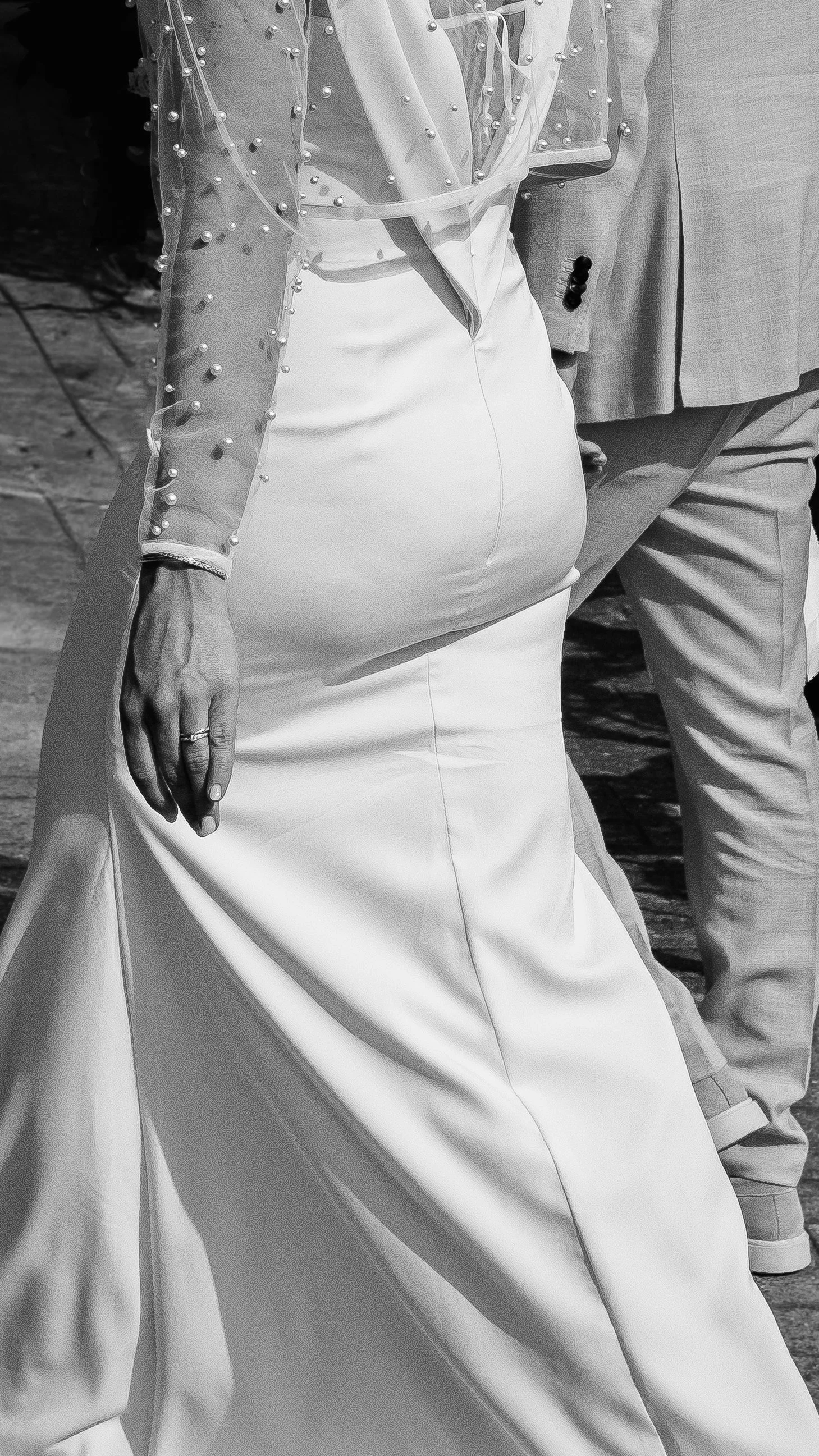Close-up of a person wearing a white dress with a long train and a sheer, sparkly top with pearl embellishments, standing outdoors.