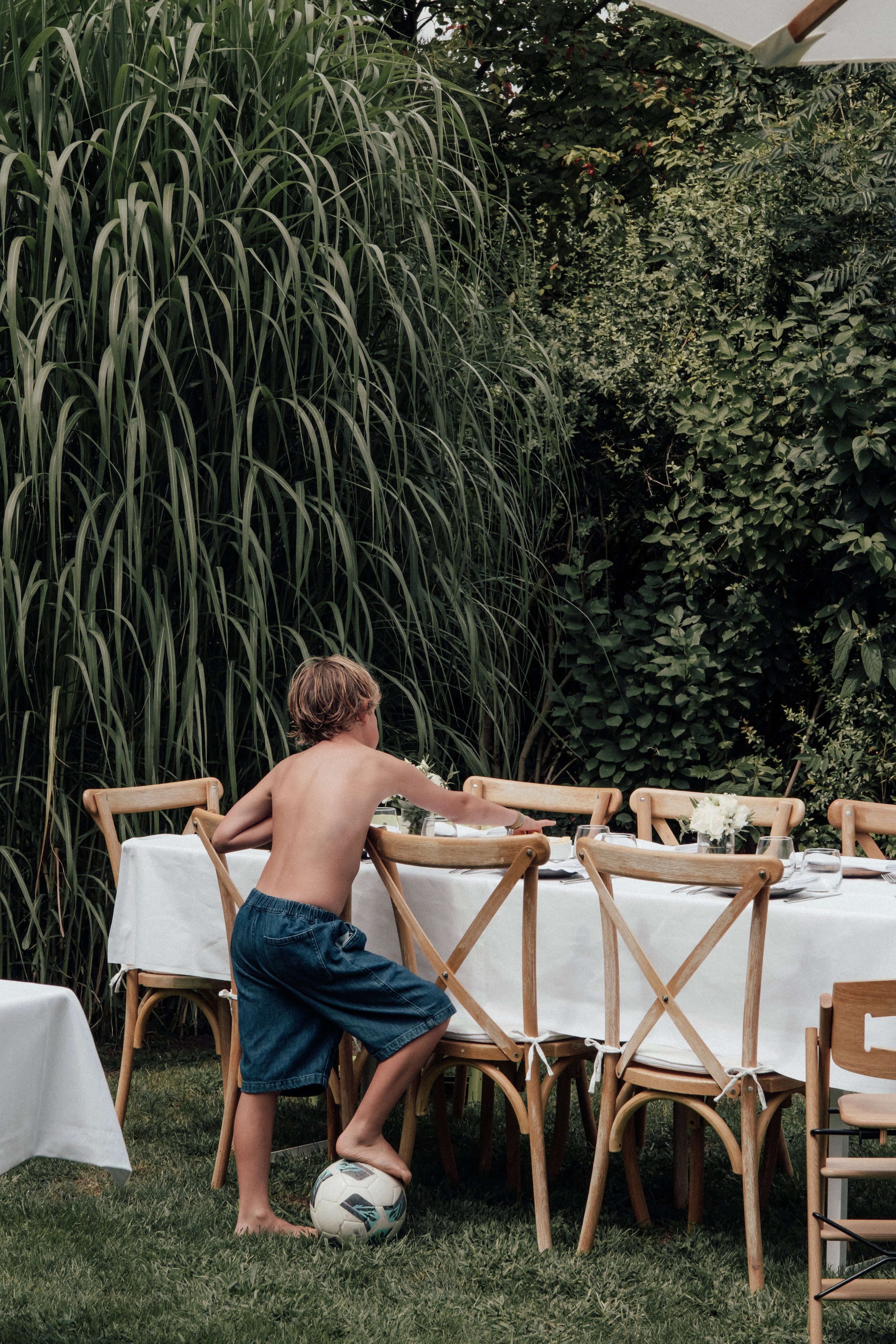 A shirtless young boy in blue shorts playing soccer next to a long outdoor dining table with chairs, set with plates and flowers, in a garden with tall green plants and bushes.
