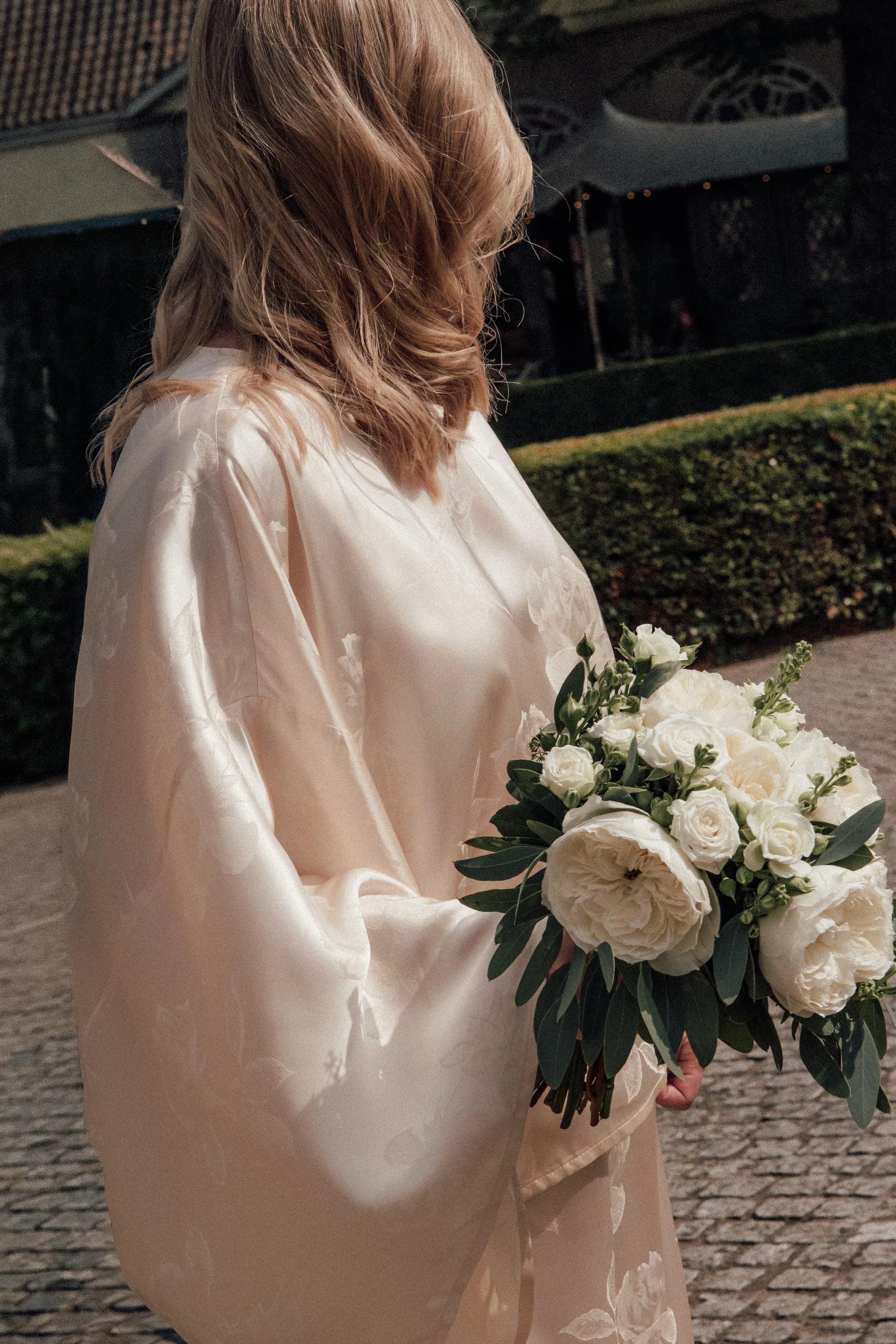A woman in a light-colored satin or silk dress holding a bouquet of white roses and greenery, standing outdoors on a cobblestone path, with a hedge and a building with large air conditioning units in the background.