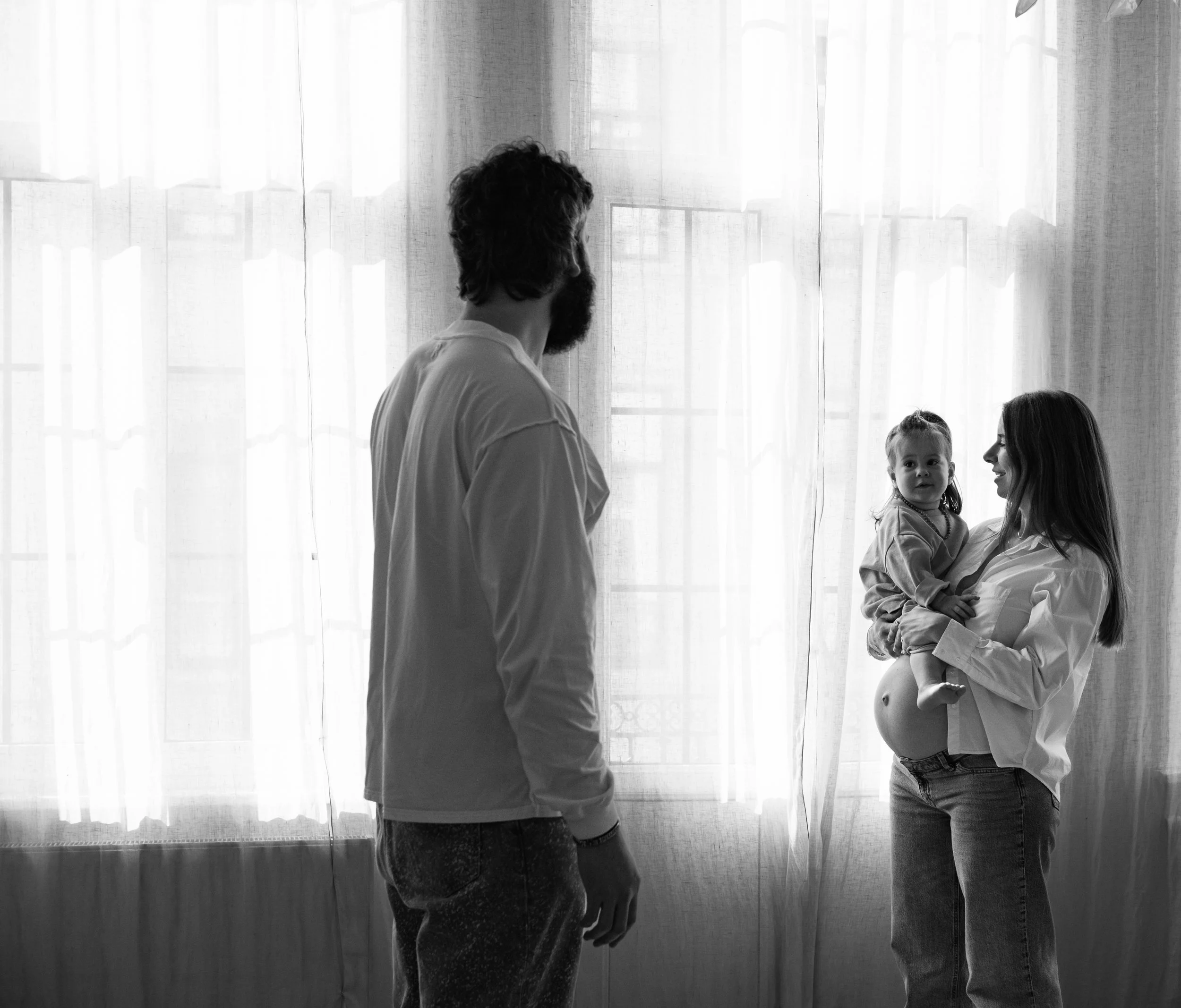 A man with dark hair and a beard looking at two young girls, one of whom is smiling while being held by a woman, against a backdrop of large windows with curtains.