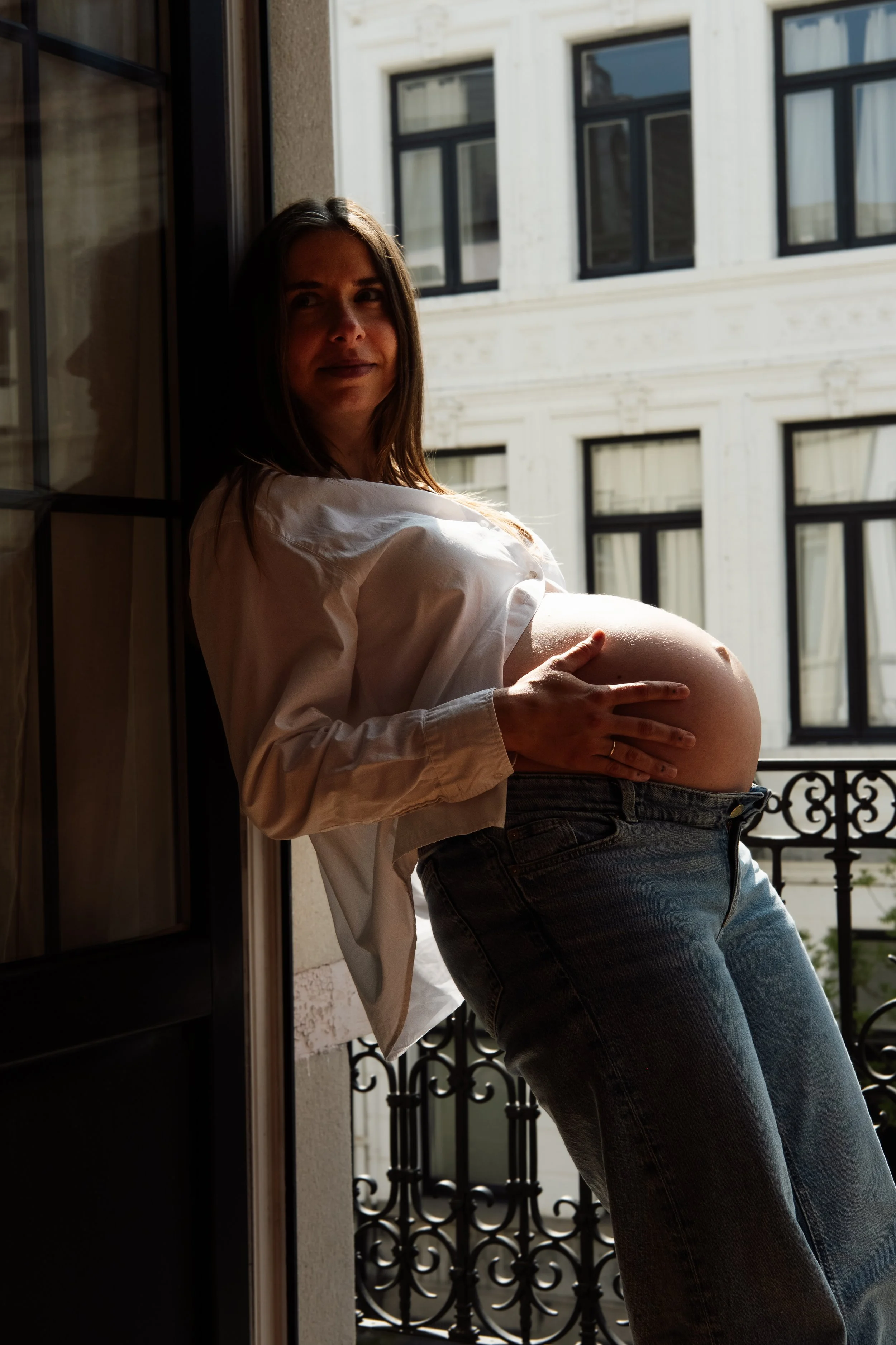 A pregnant woman standing on a balcony, touching her belly, with a building and windows in the background.
