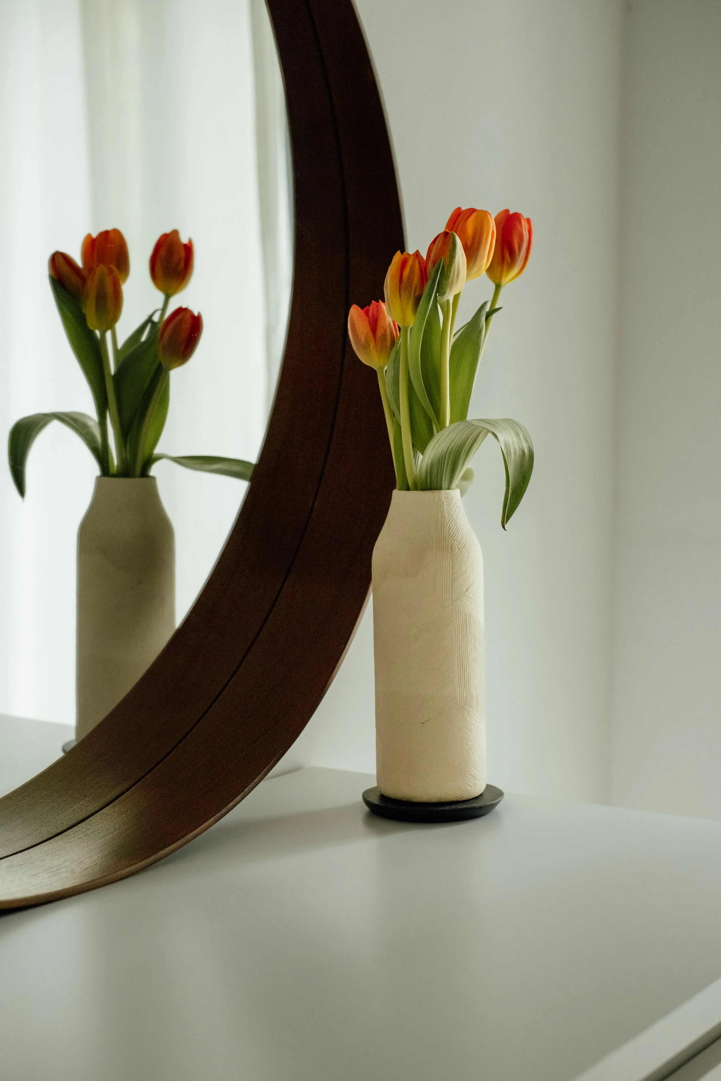 A white vase holding orange tulips with green leaves, reflected in a large mirror, on a white surface against a light-colored wall.