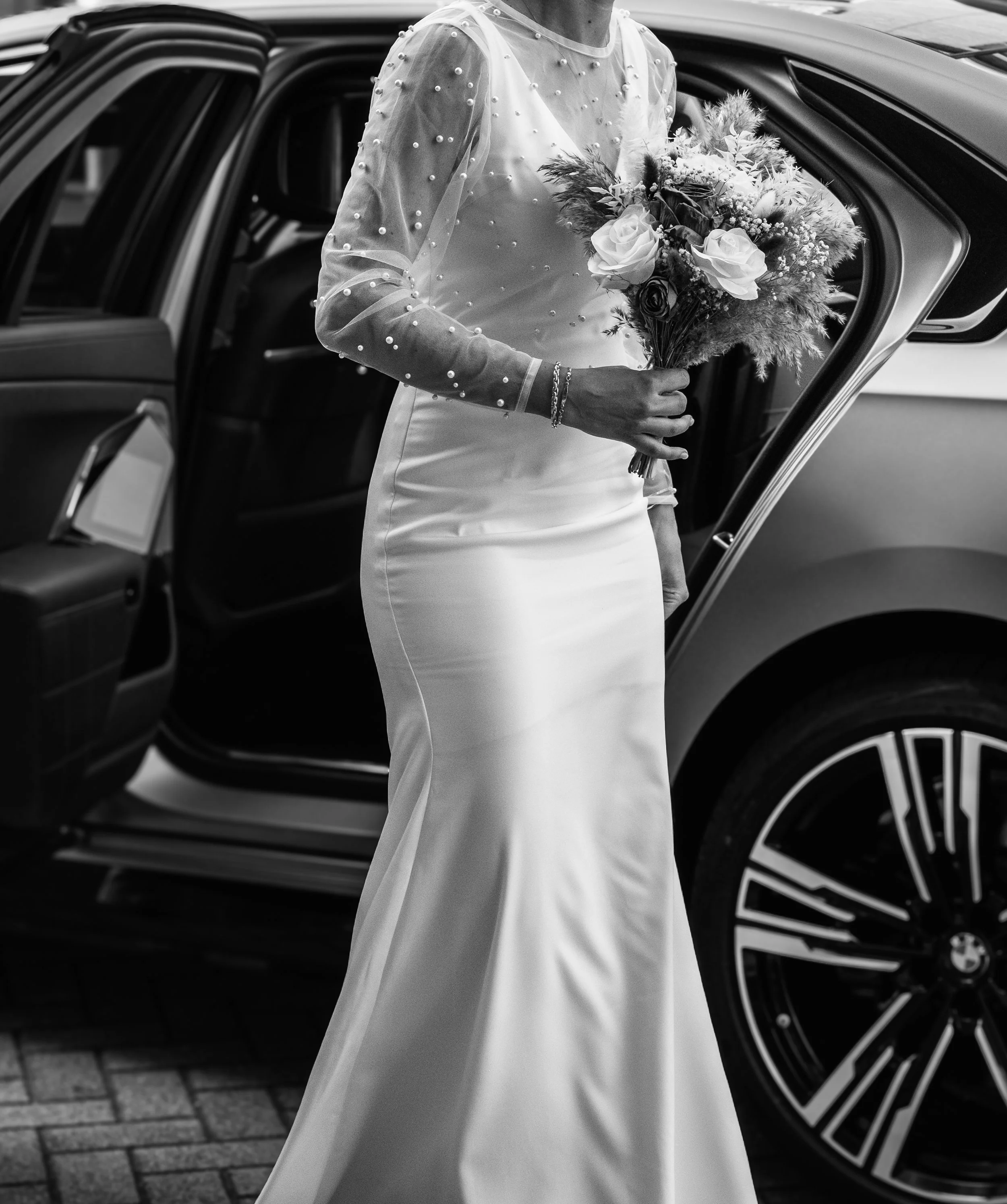 A bride in a elegant gown holding a bouquet of roses and flowers, standing next to a luxury car with an open door.