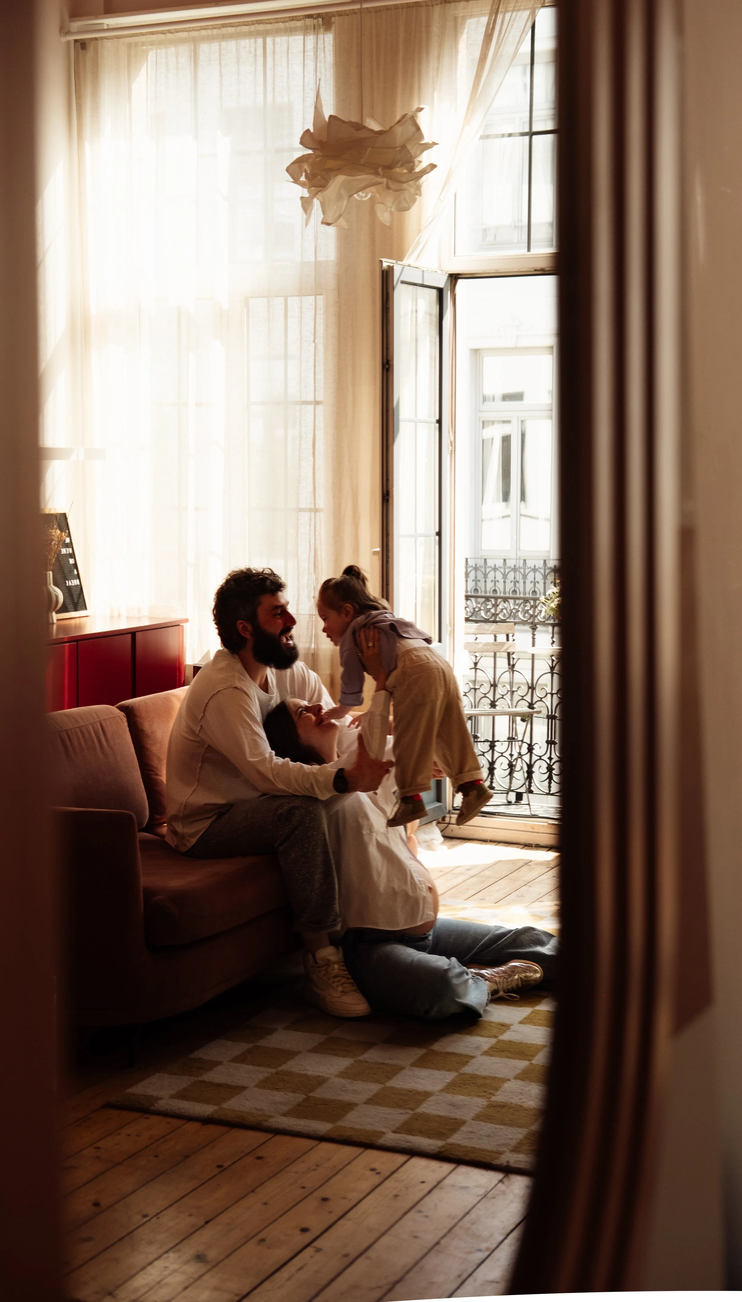 A family enjoying quality time together in a living room with large windows and open balcony doors, allowing natural sunlight to fill the space.
