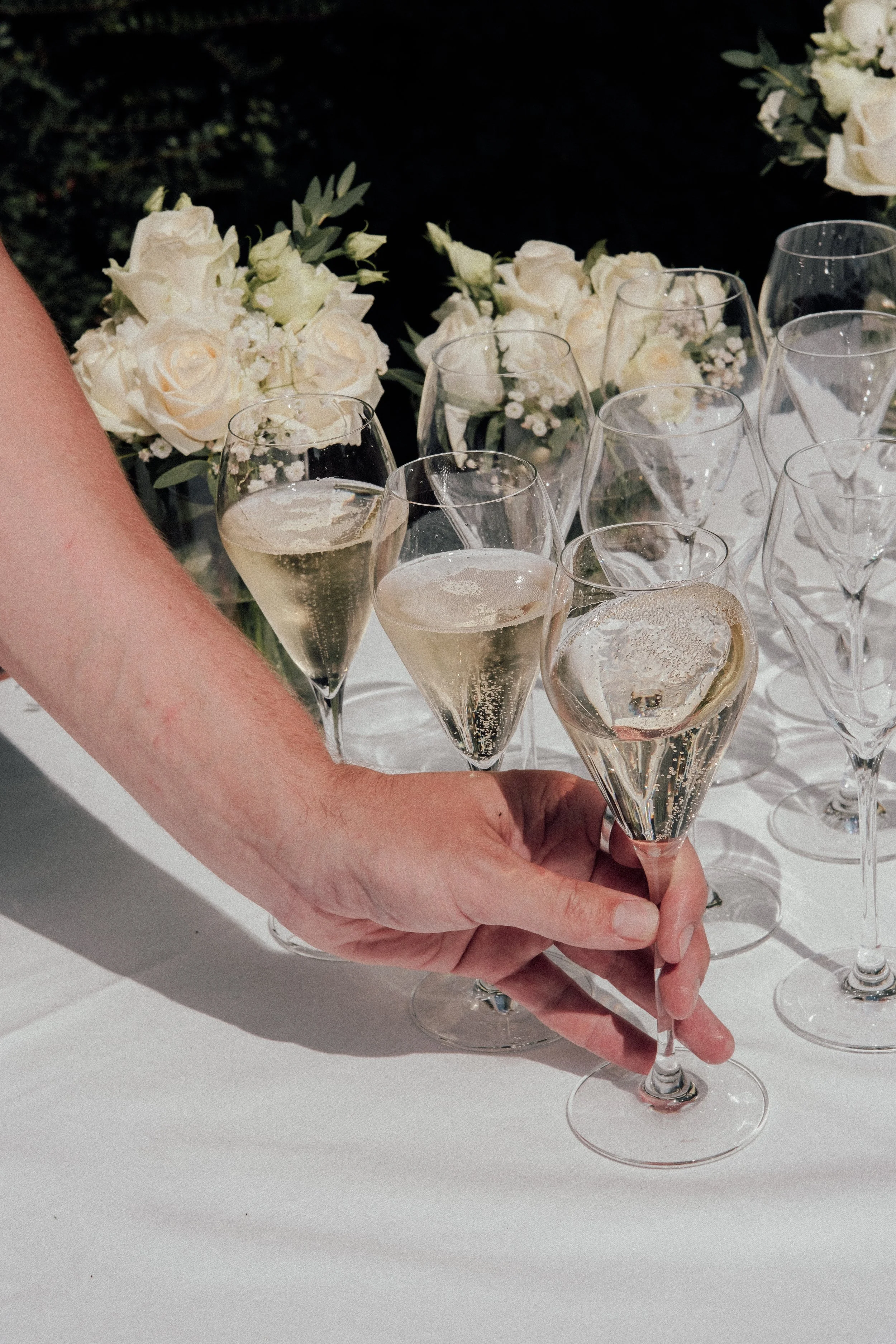 Person holding several glasses of champagne at a celebration table with white floral arrangements.