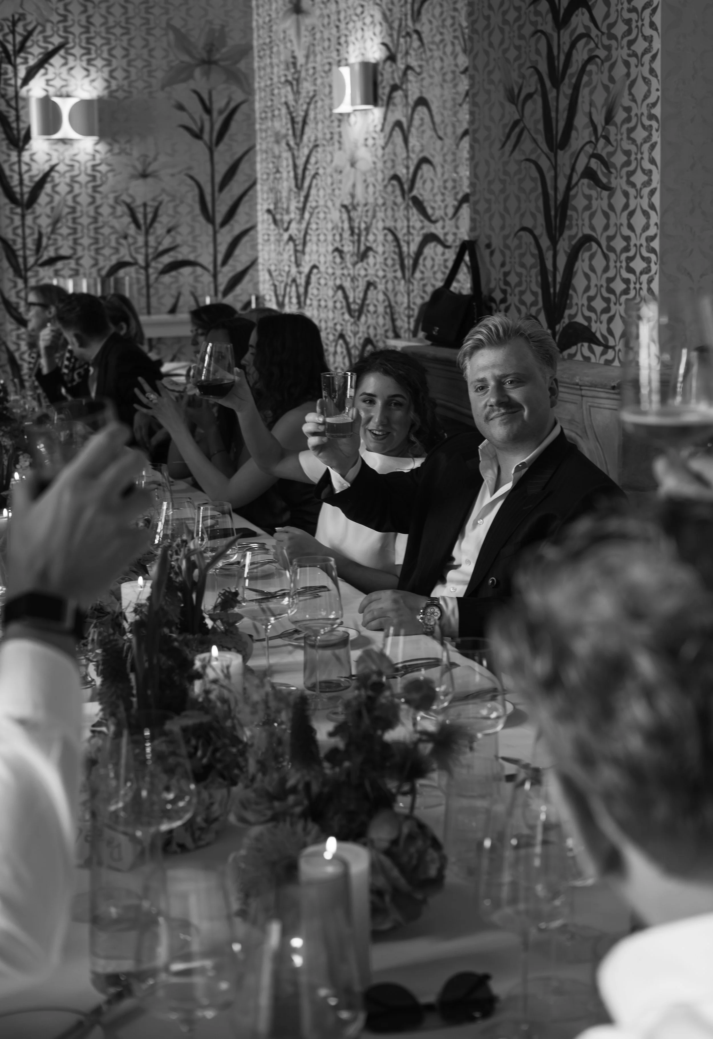 People sitting at a banquet table during a celebration, raising glasses for a toast, with floral centerpieces and decorative wallpaper in the background.