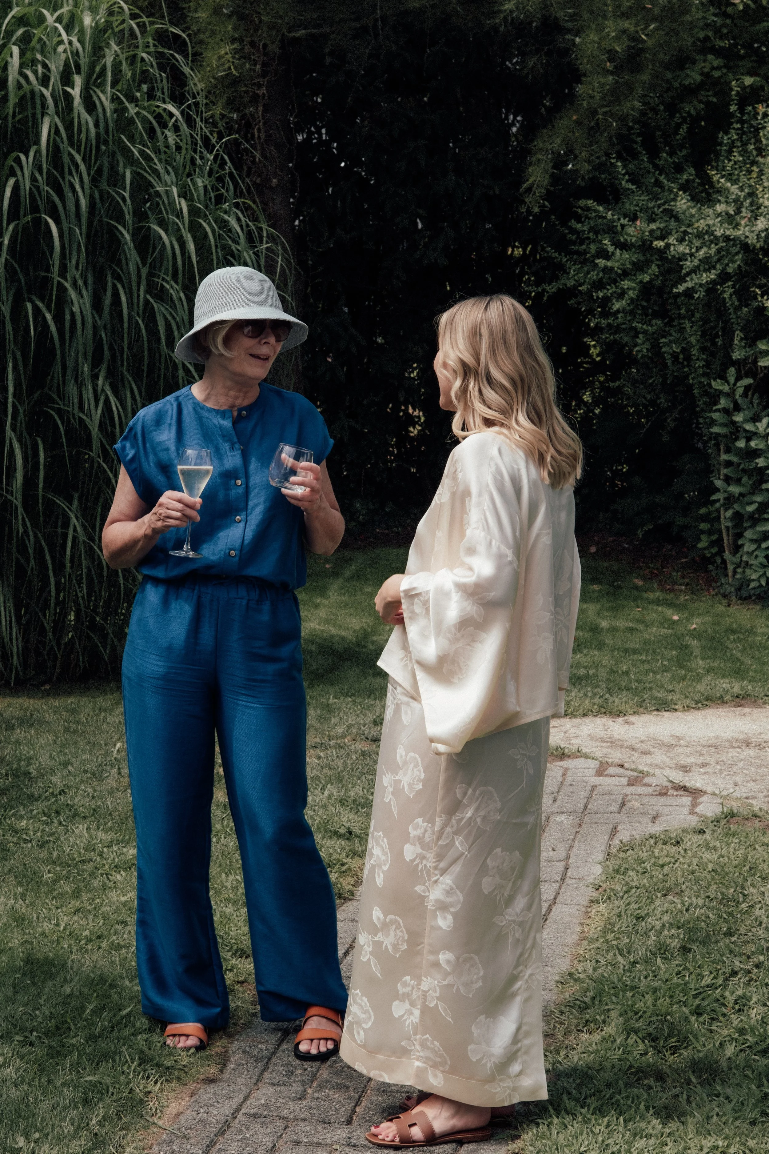 Two women having a conversation outdoors at night, one wearing a white hat, sunglasses, and a blue jumpsuit holding wine glasses, the other in a cream floral kimono and sandals, standing on a brick sidewalk near grass and trees.