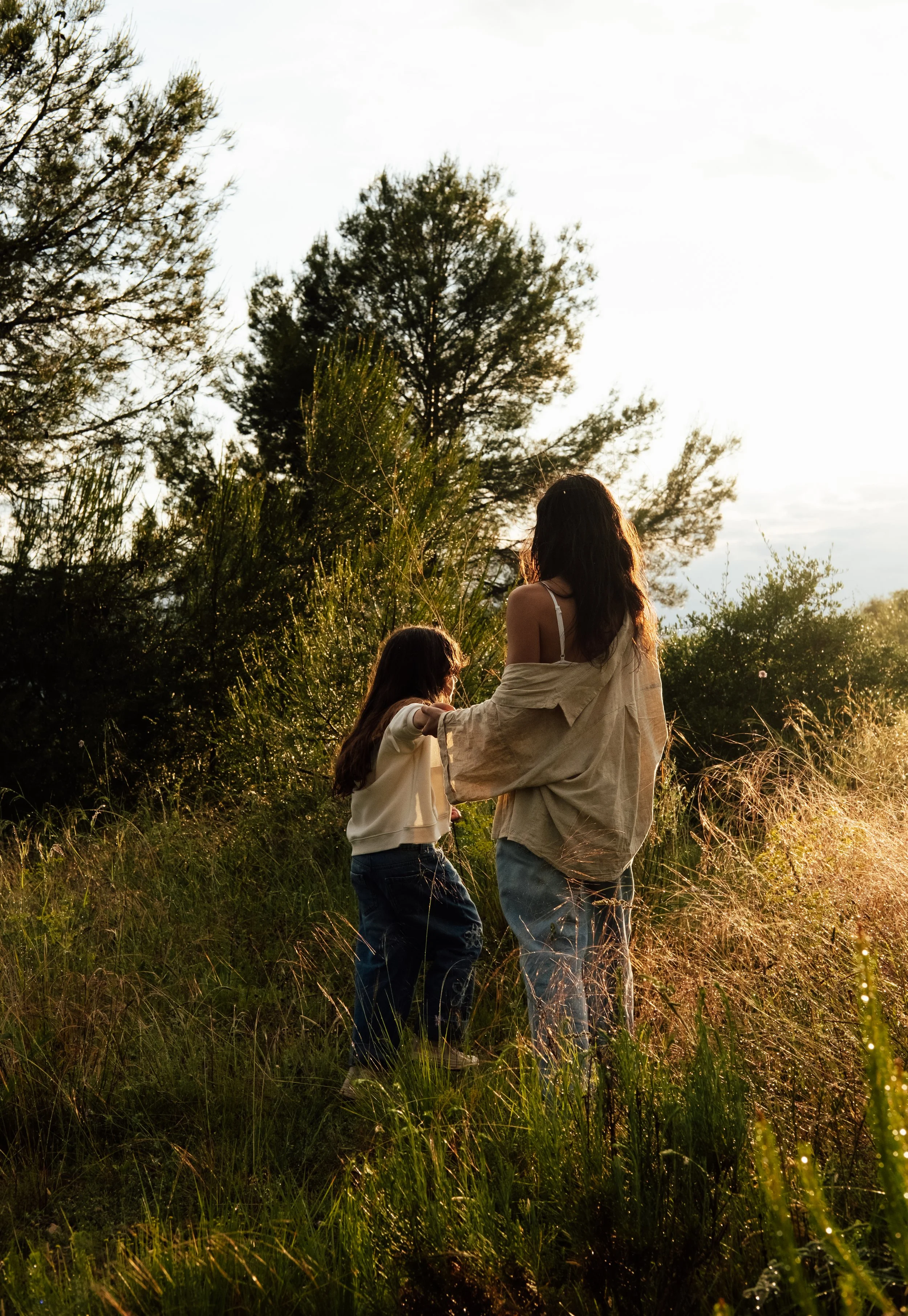 A woman and a girl standing in a grassy field with trees in the background during sunset, holding hands.
