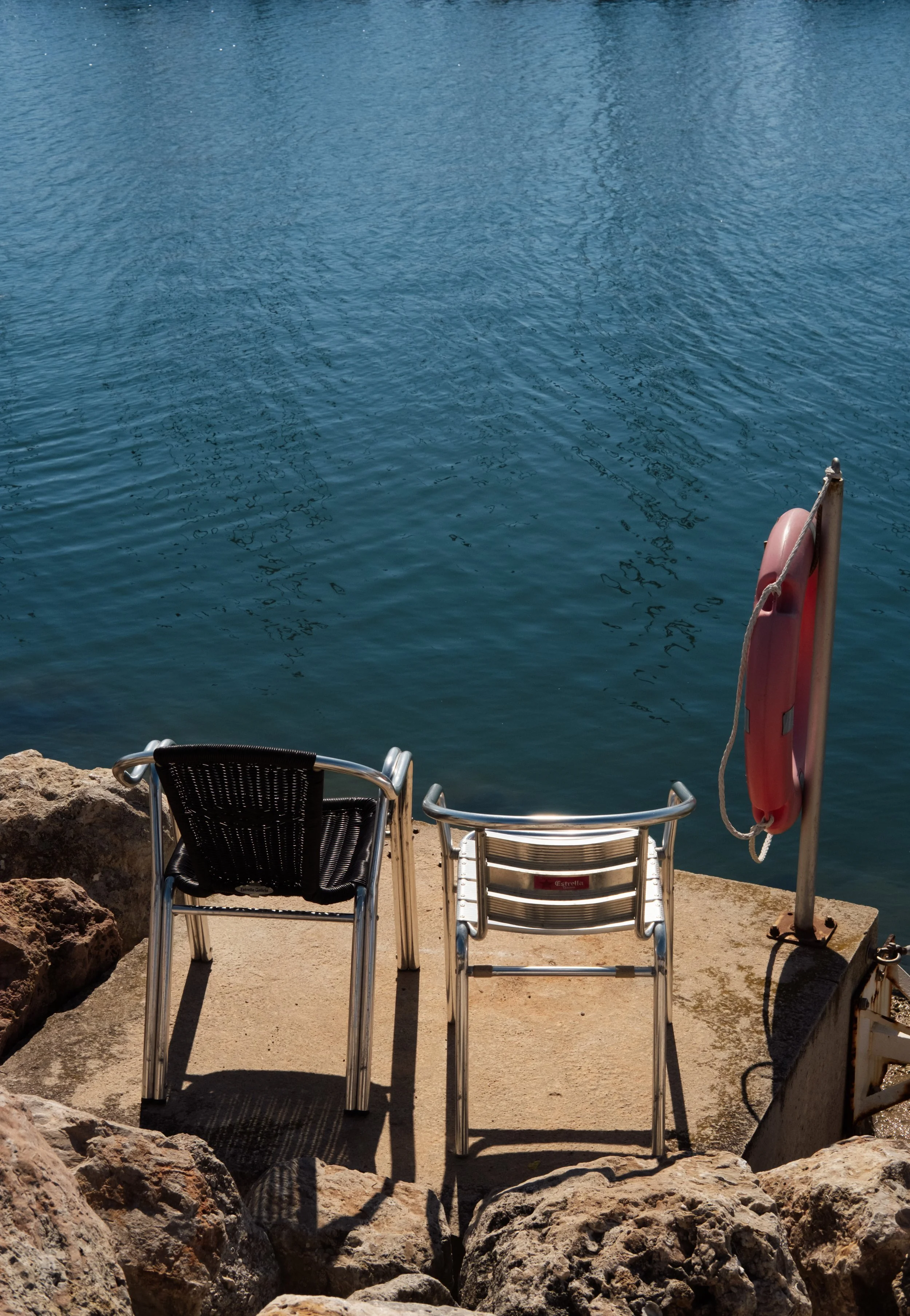 Two chairs with black and silver metal frames on a concrete surface overlooking a body of water. A orange life preserver is attached to a metal pole on the right side of the image. Rocks surround the seating area.