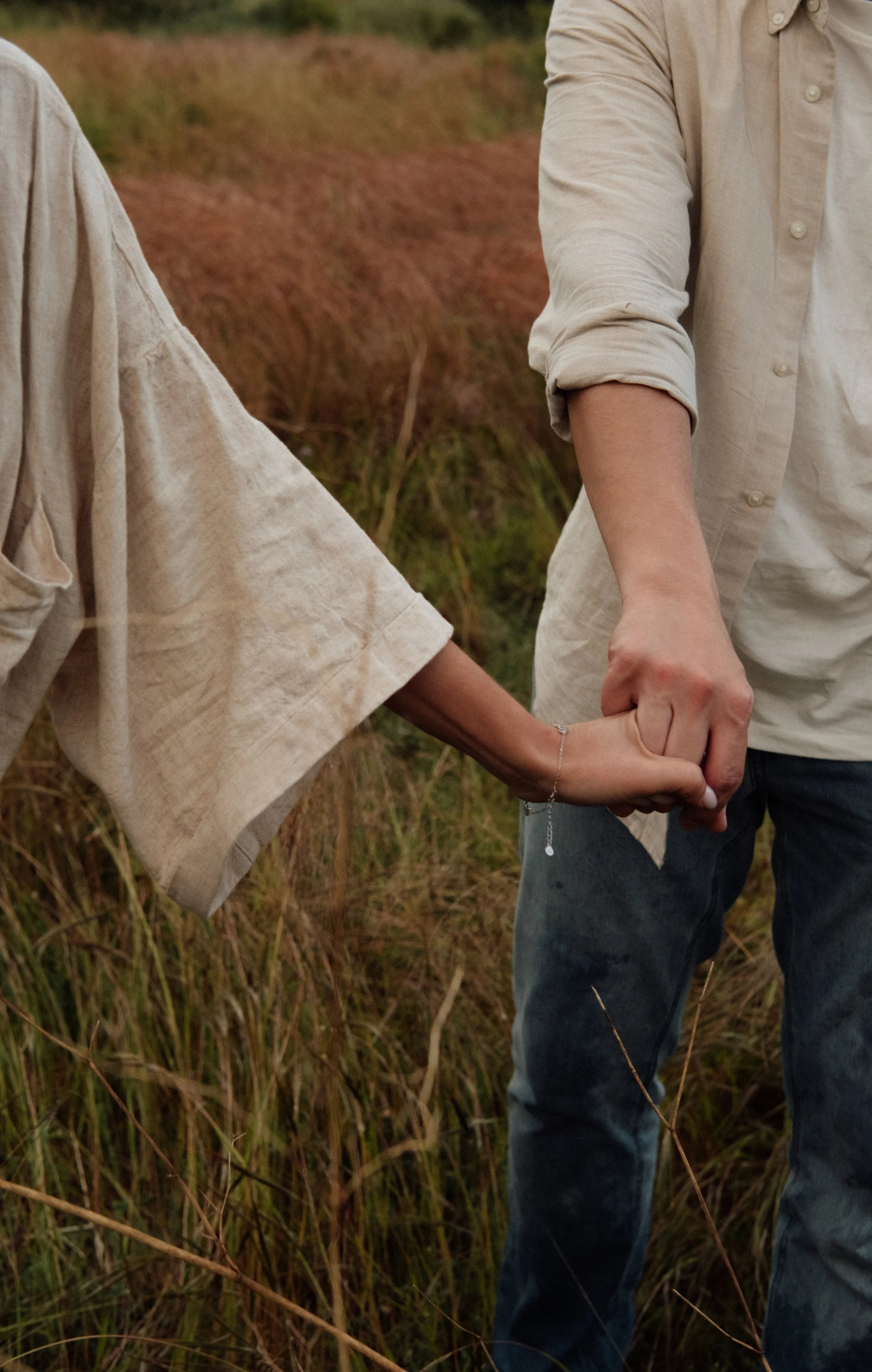 A man and woman holding hands outdoors in a field with tall grass, wearing beige and denim clothing.