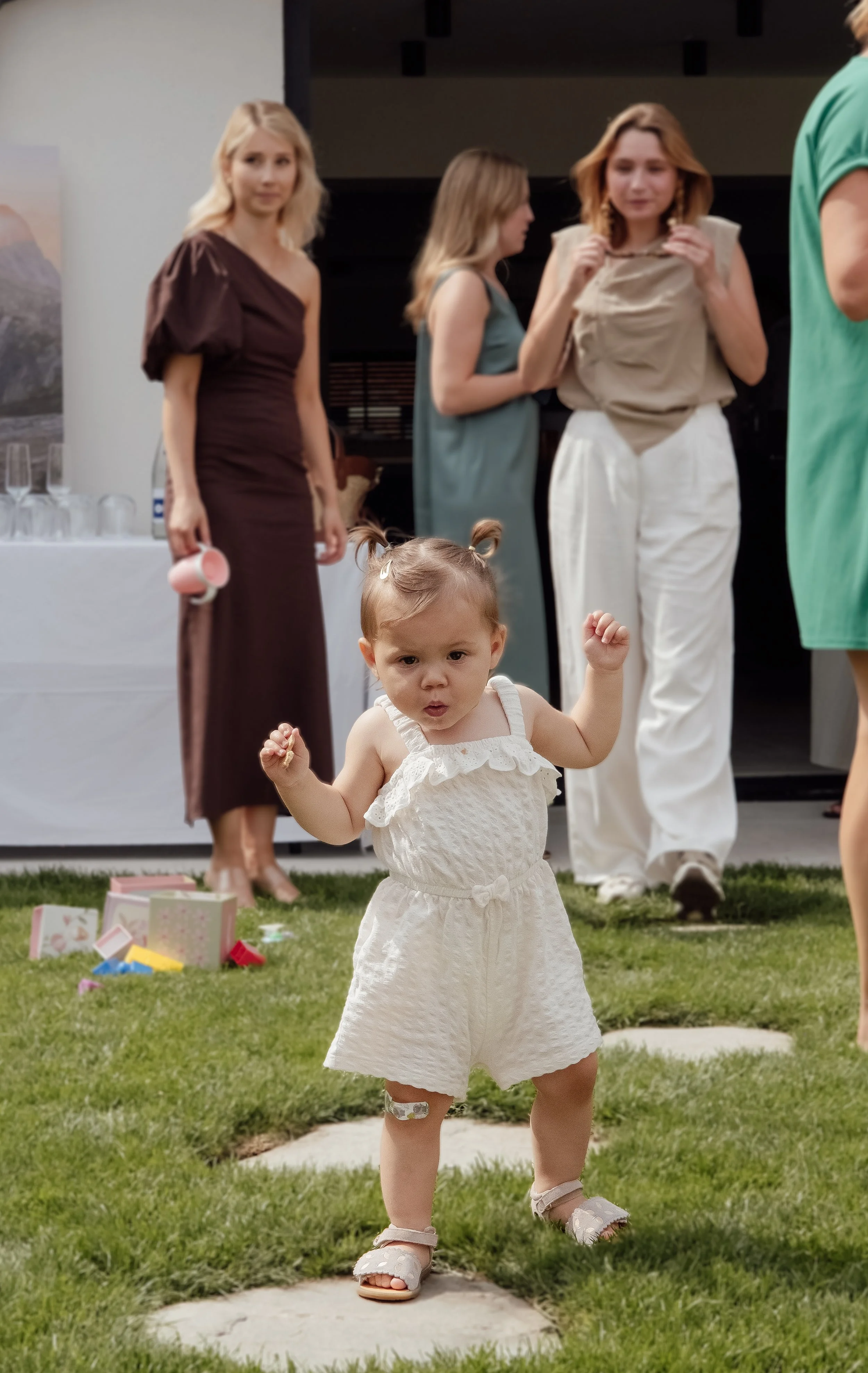 A young girl with short hair in a white dress standing on grass with her fist raised, in front of a group of women at a gathering or party.