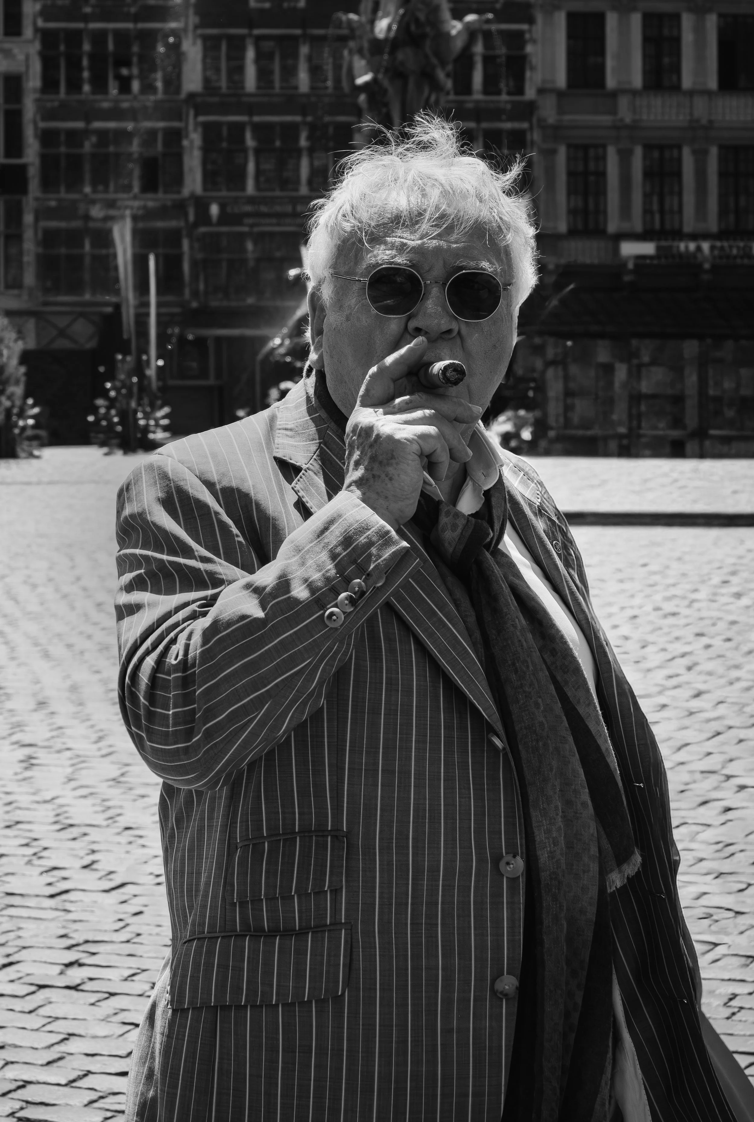 A black and white photo of an older man with white hair wearing round sunglasses, a striped blazer, and a scarf, smoking a cigar in an urban square with cobblestone pavement and buildings in the background.